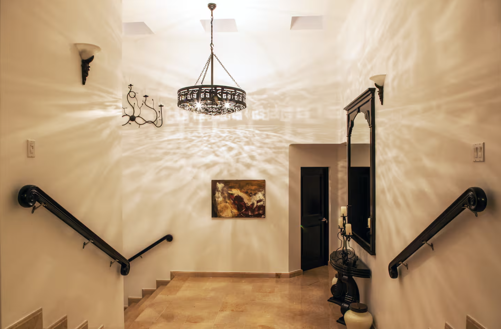 Elegant staircase landing with warm lighting, wall sconces, chandelier, artwork, mirror, and decorative table with candles.