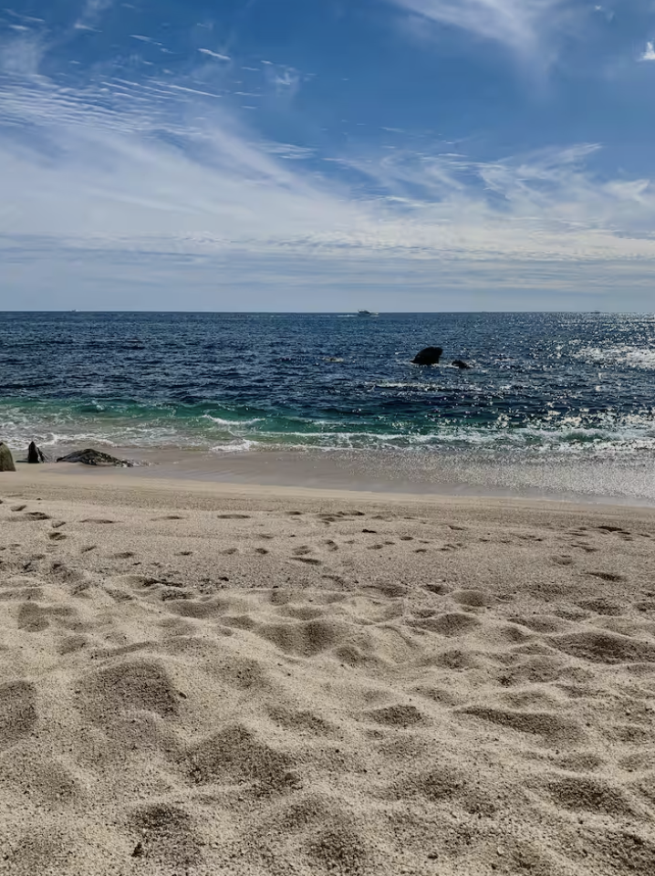 View of a sandy beach with the ocean and sky in the background, some rocks near the shore, and a ship visible on the water.
