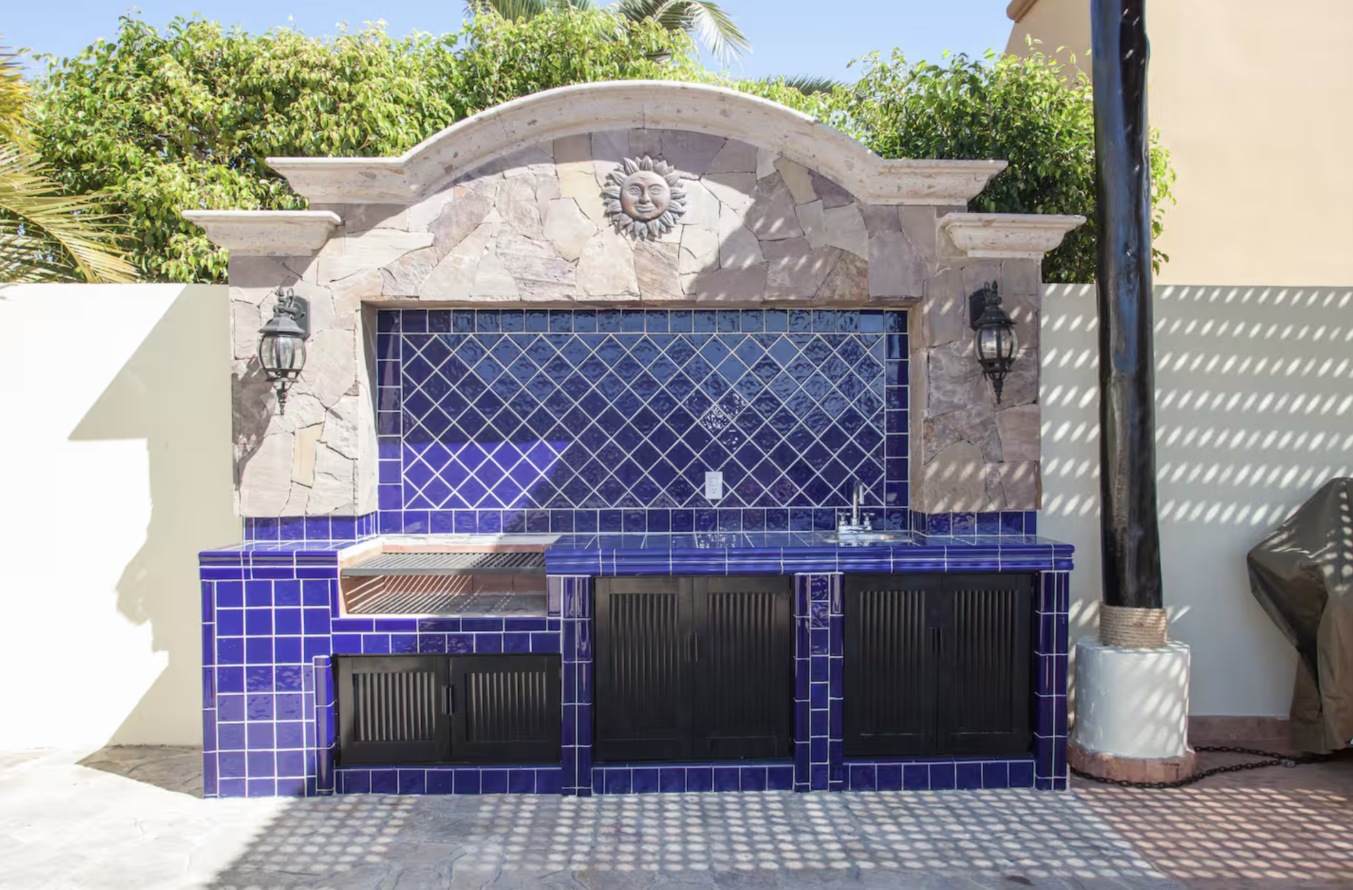 Outdoor kitchen with blue tile backsplash, black cabinets, and stone arch with sun and moon face decoration, surrounded by greenery.