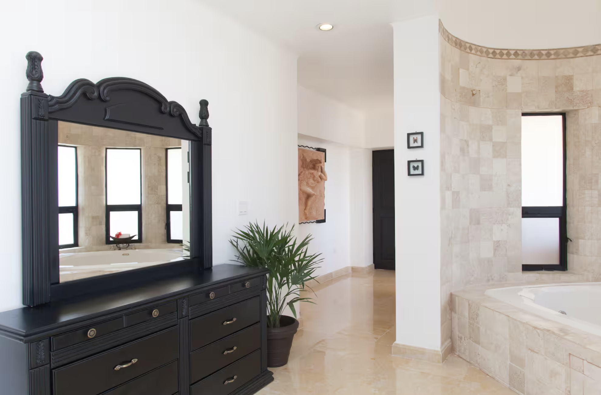 Indoor bathroom with black dresser, large mirror, potted plant, artwork, and a bathtub near windows.