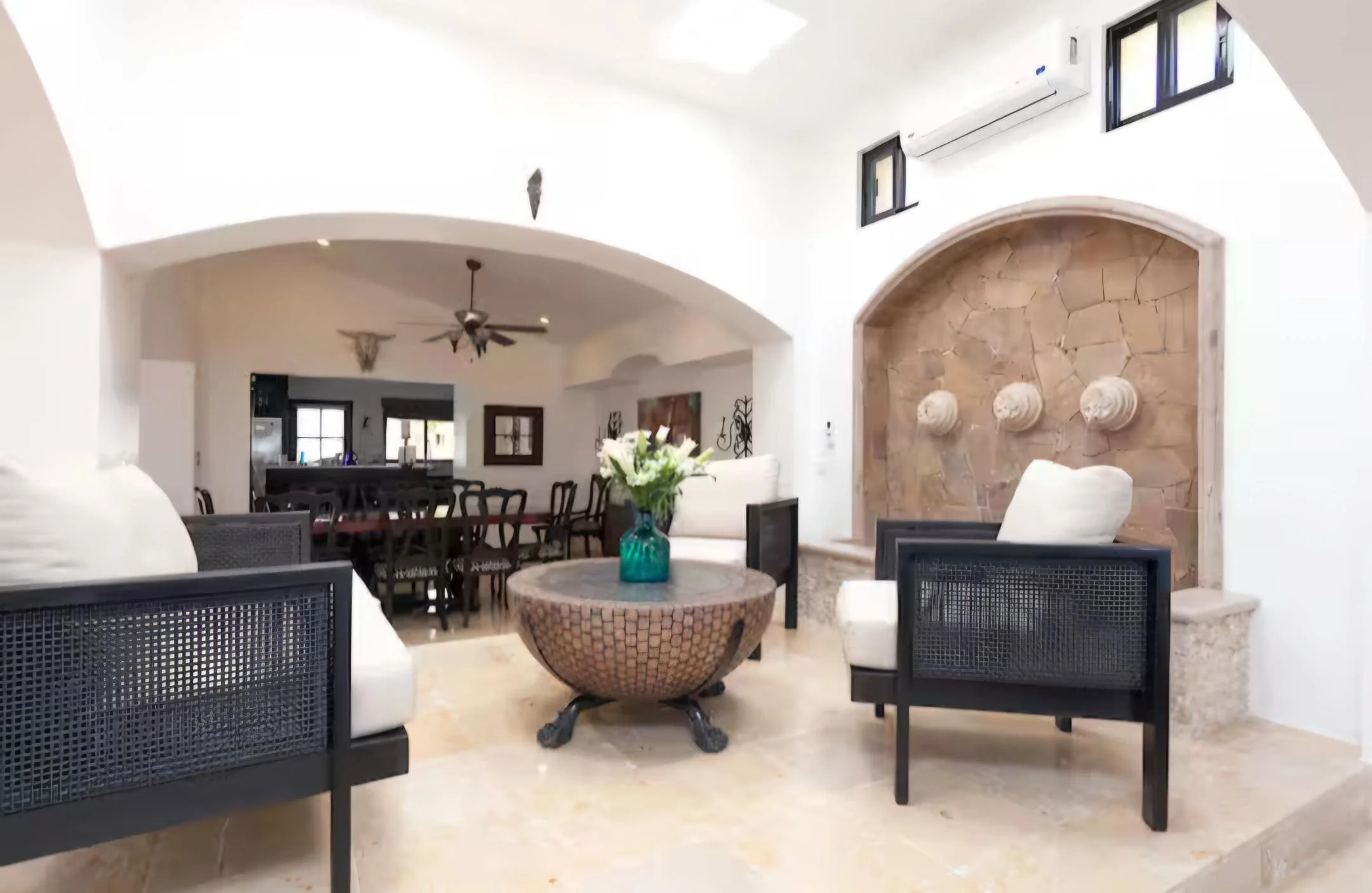 Living room with white walls, arched ceiling, black furniture including chairs and a table with a vase of white flowers, and a decorative stone wall with three wall-mounted pottery pieces. The Marlin Hunter luxury villas in Cabo