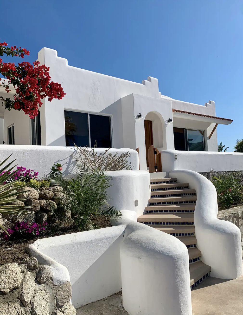 A white Mediterranean-style house with stairs leading up to the entrance, decorated with vibrant pink and red flowering plants and green foliage, under a clear blue sky.