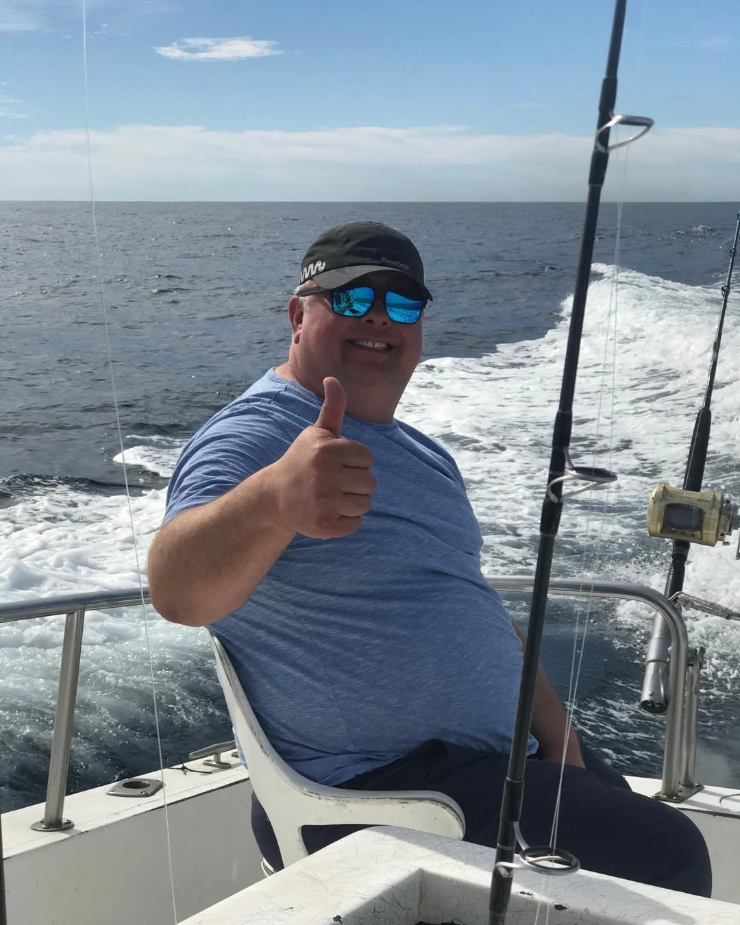 A man sitting on a boat at sea, smiling and giving a thumbs-up, with fishing rods nearby, under a partly cloudy sky. The Marlin Hunter Fishing Charter Trip
