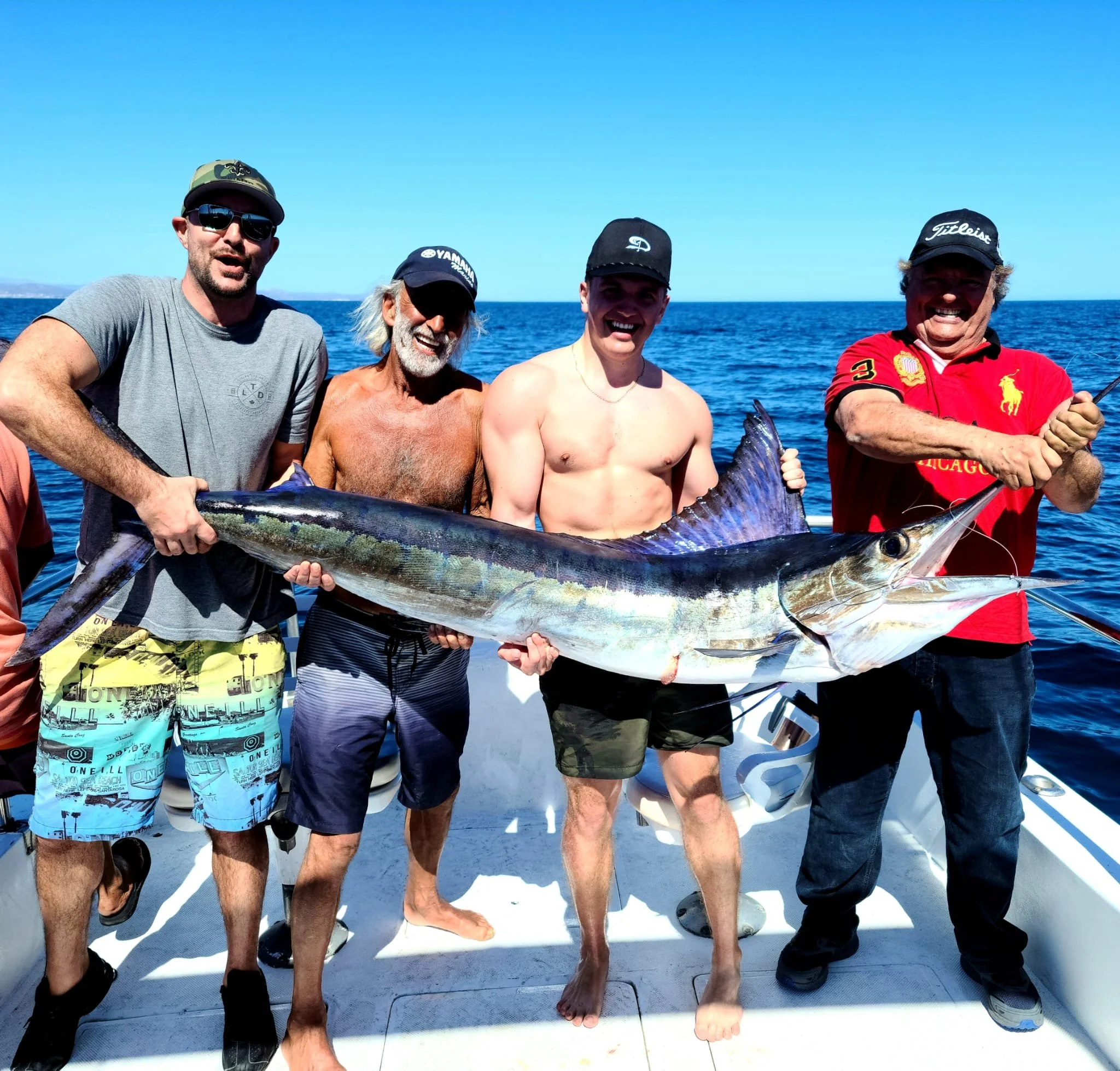 Group of four men on a boat holding a large fish, possibly a marlin, after a successful fishing trip. The ocean and clear blue sky are in the background.