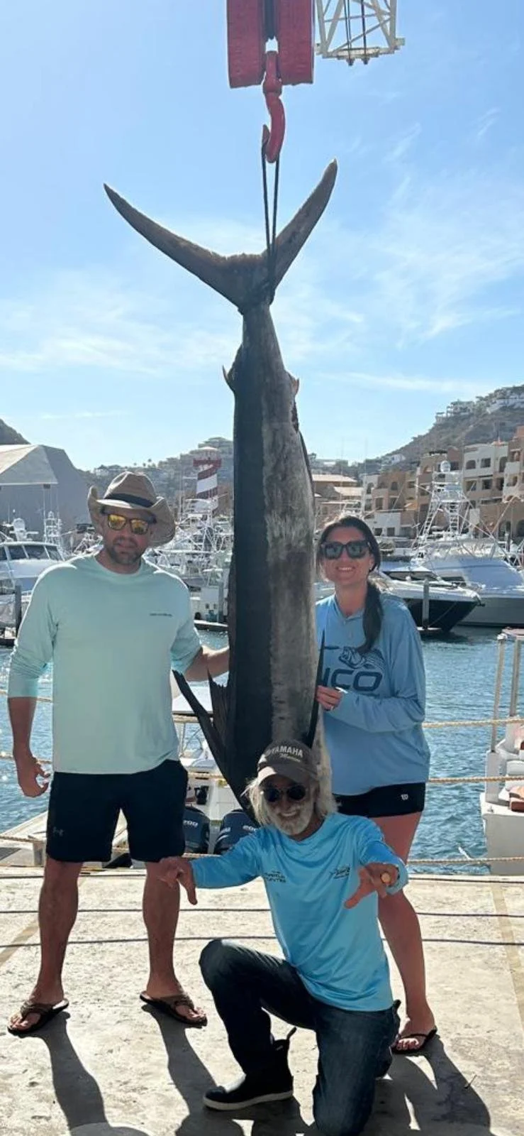 Three people in casual clothing and sunglasses stand on a dock by the harbor, holding a large fish with a whale tail, which is suspended from a crane. The background features boats, a marina, and coastal buildings under a blue sky.