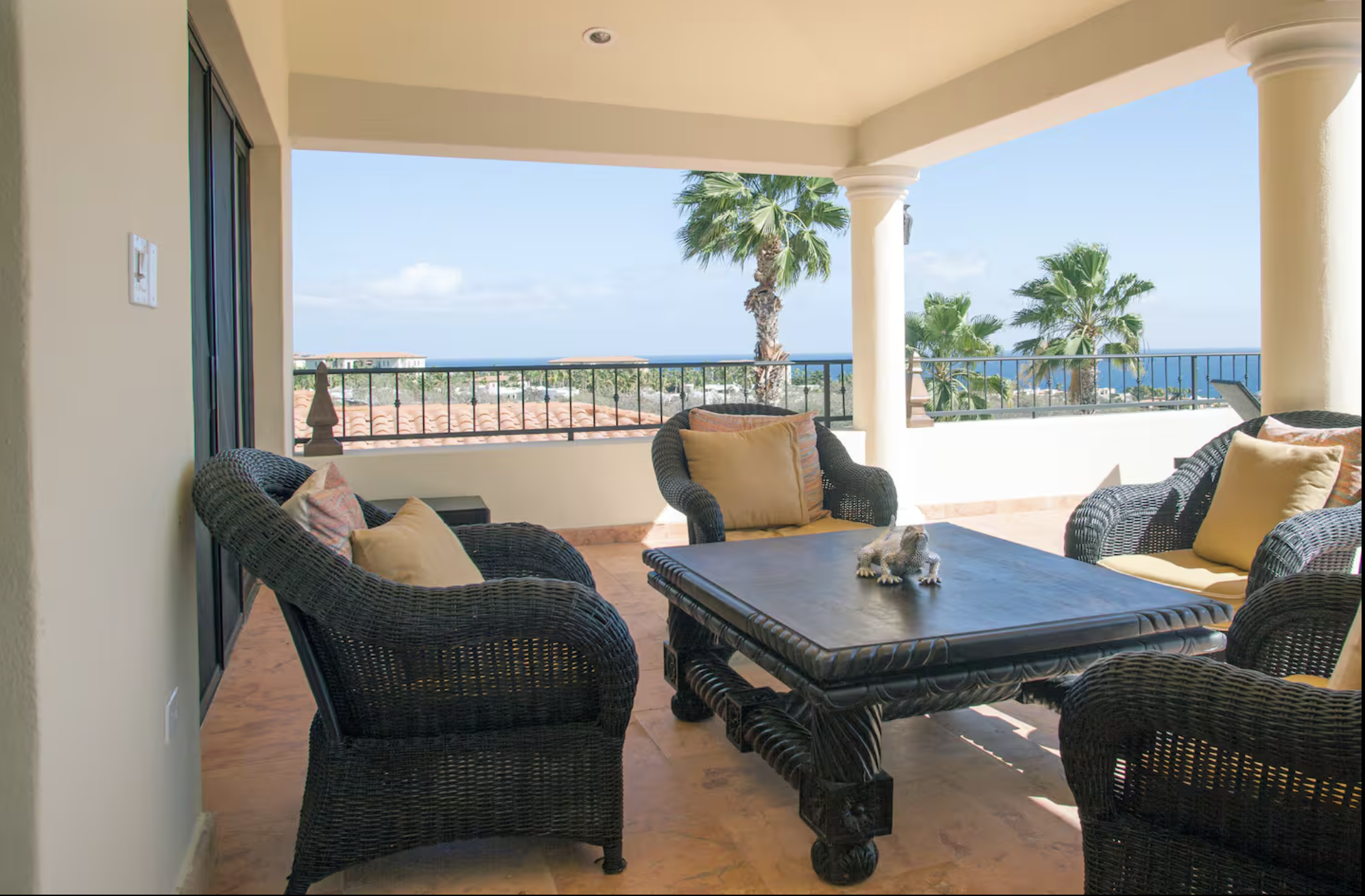 Outdoor balcony with wicker furniture, a table, and yellow cushions, overlooking palm trees, rooftops, and the ocean under a blue sky.