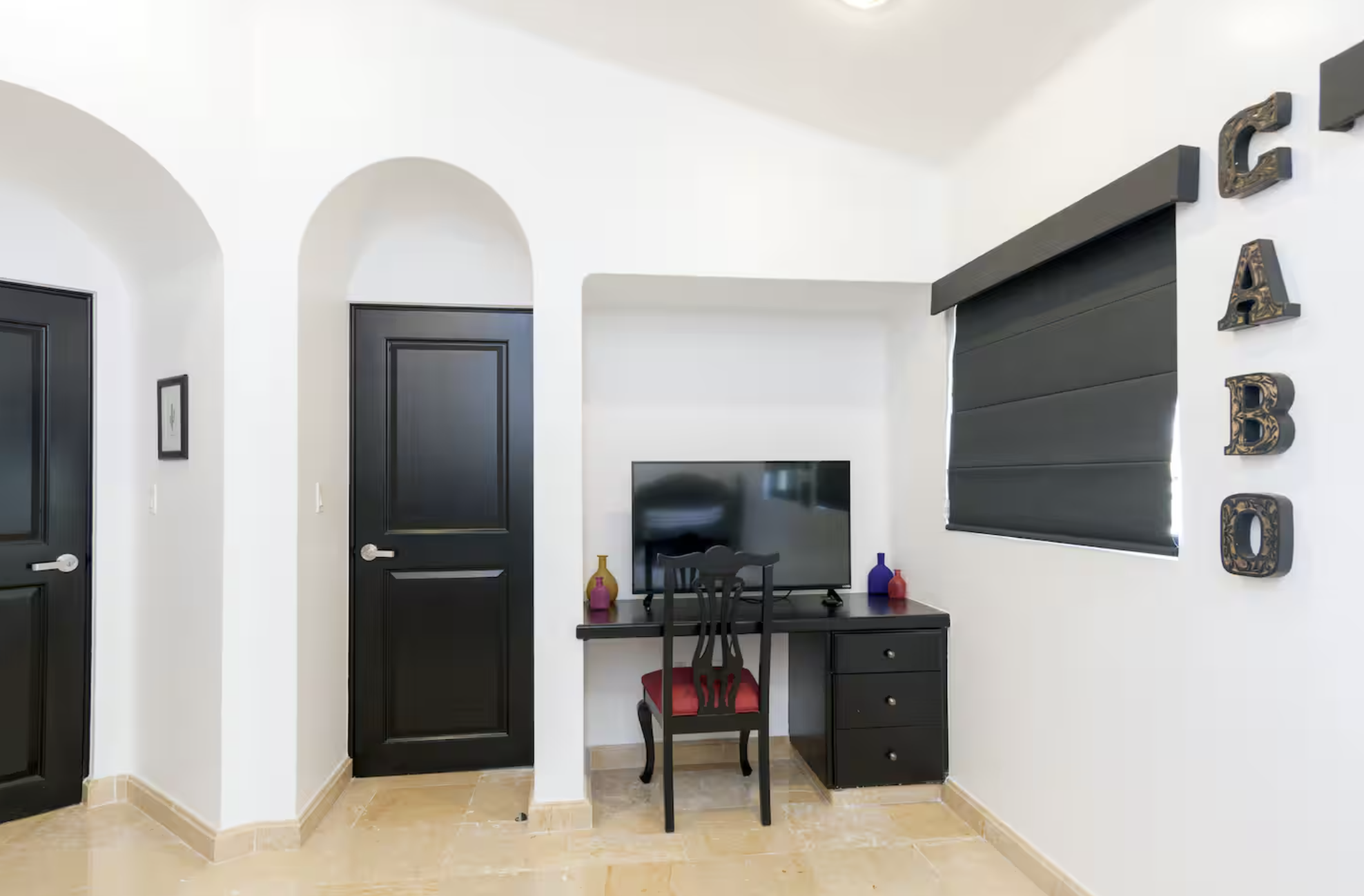 Living room corner with a black desk, computer monitor, black chair with red cushion, three vases in yellow, purple, and red on the desk, black windows with blinds, white walls, and black doors, with the word 'CABO' vertically on the wall.