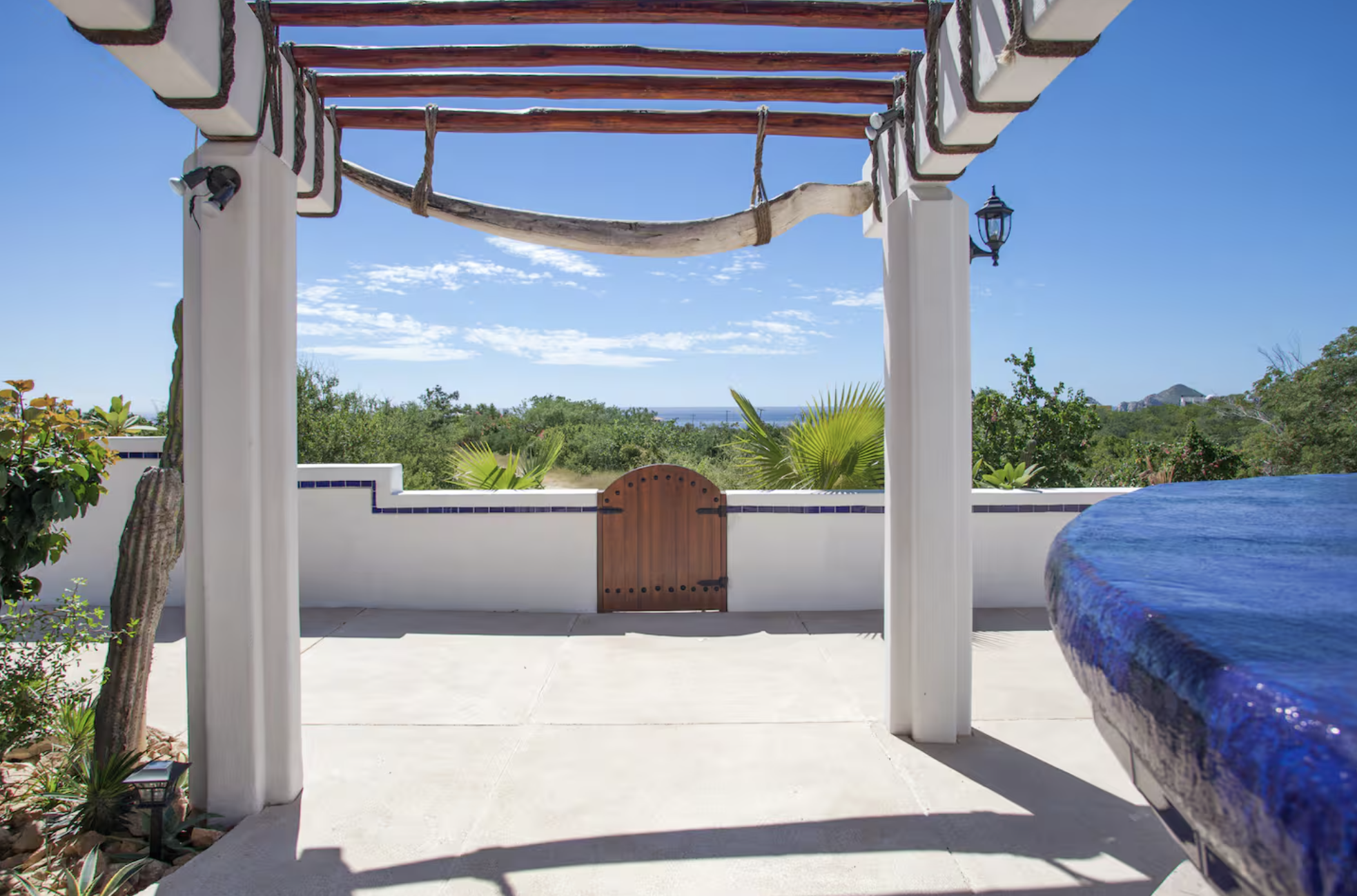 View of an outdoor patio with a white wall and small wooden gate, tropical plants, and a blue sky with a few clouds.