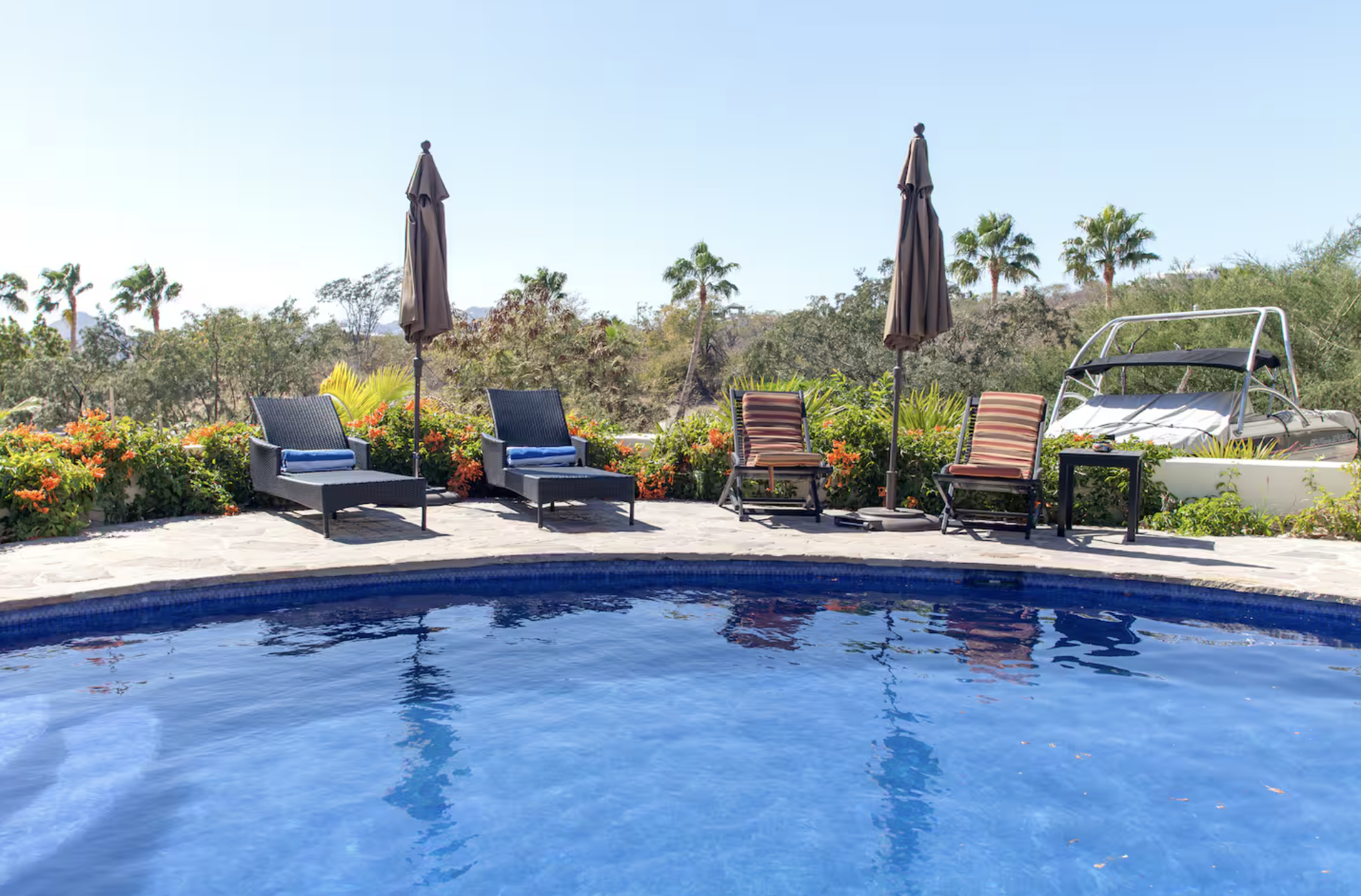 A backyard scene with a swimming pool in the foreground, surrounded by chairs, umbrellas, and lush greenery, with palm trees and a boat in the background.