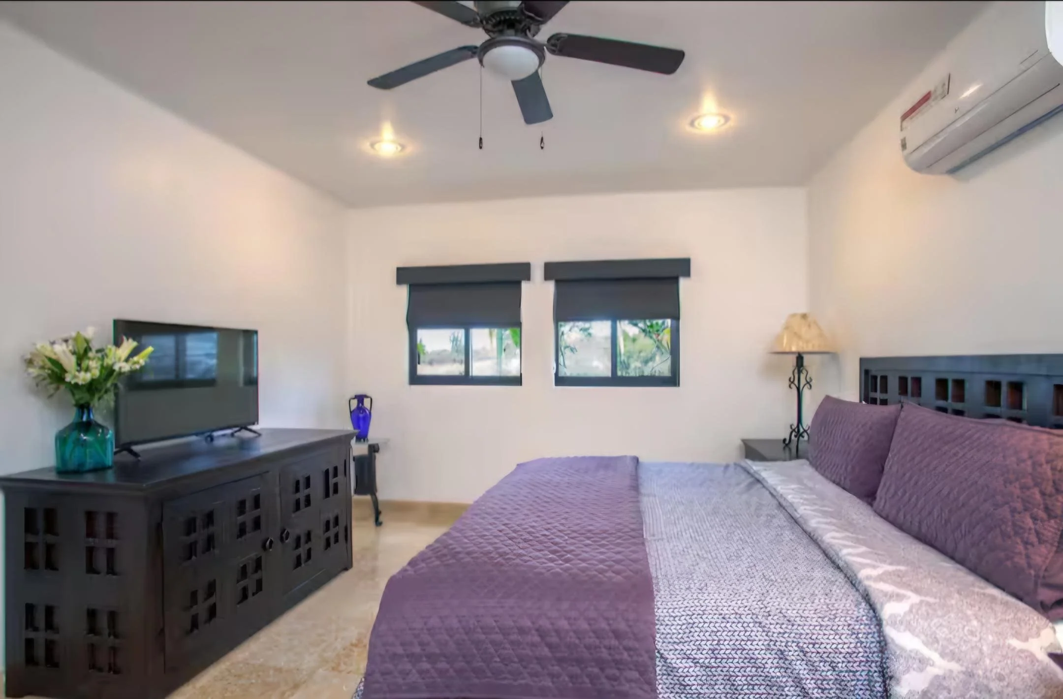 A bedroom with two small windows, a queen-sized bed with purple bedding, a nightstand with a lamp, a TV on a dark wood cabinet, a vase with flowers, and a ceiling fan. The Marlin Hunter luxury villas in Cabo