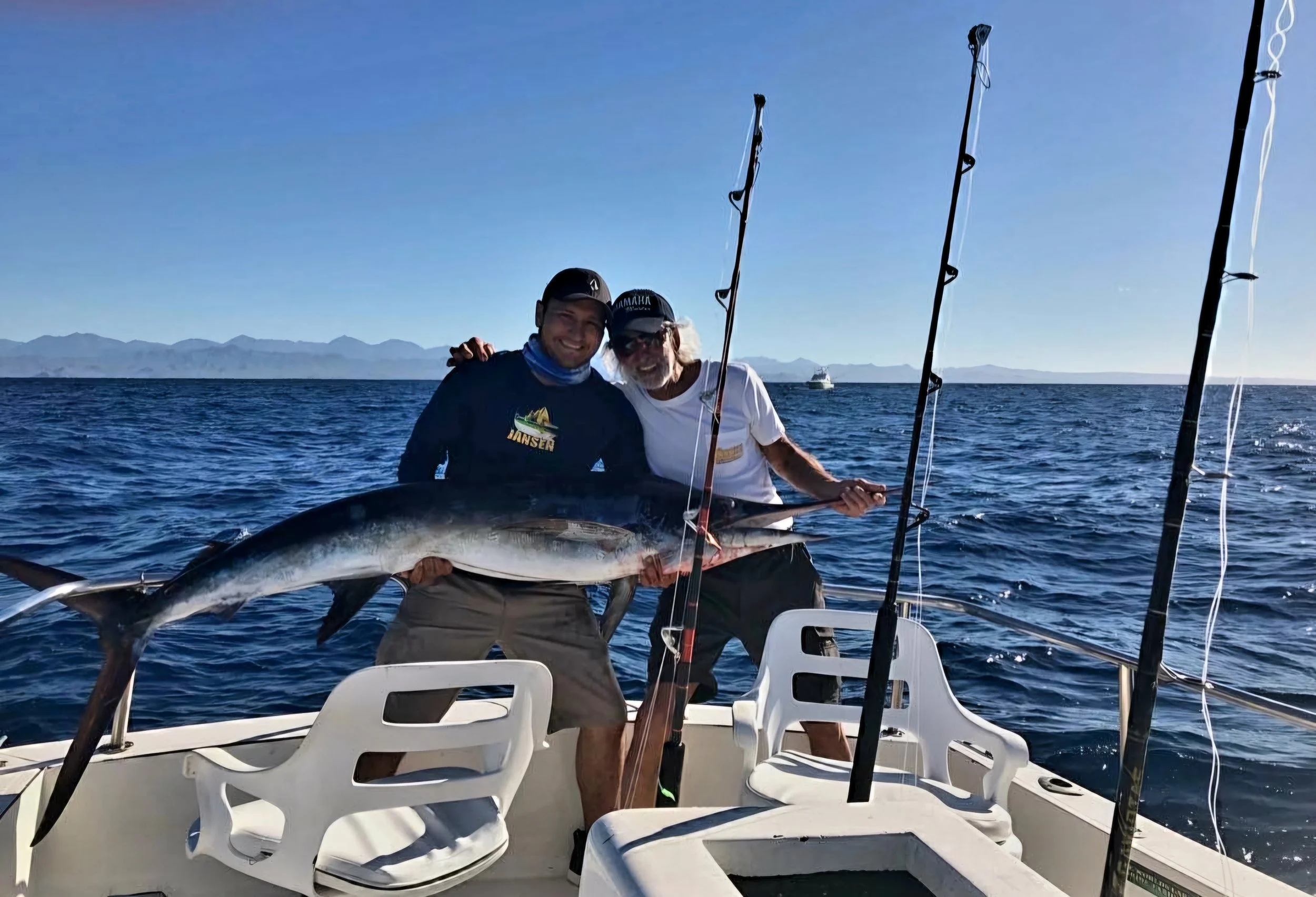 Two men on a boat holding a large fish, with fishing rods, on the open sea with mountains in the background. | The Marlin Hunter Fishing Charter Day Trip from Cabo