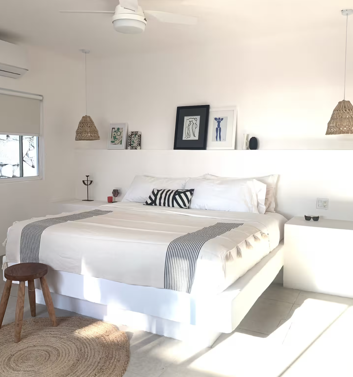 A modern bedroom with a white bed, black and white striped pillow, floating shelf with framed artwork, wicker pendant lights, and a small wooden stool on a round jute rug.
