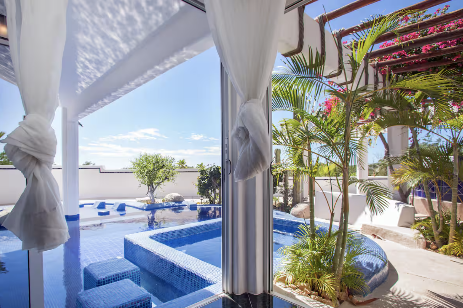 A view of a courtyard with a hot tub, lush tropical plants, and a clear blue sky, visible from inside through an open sliding door with white curtains.