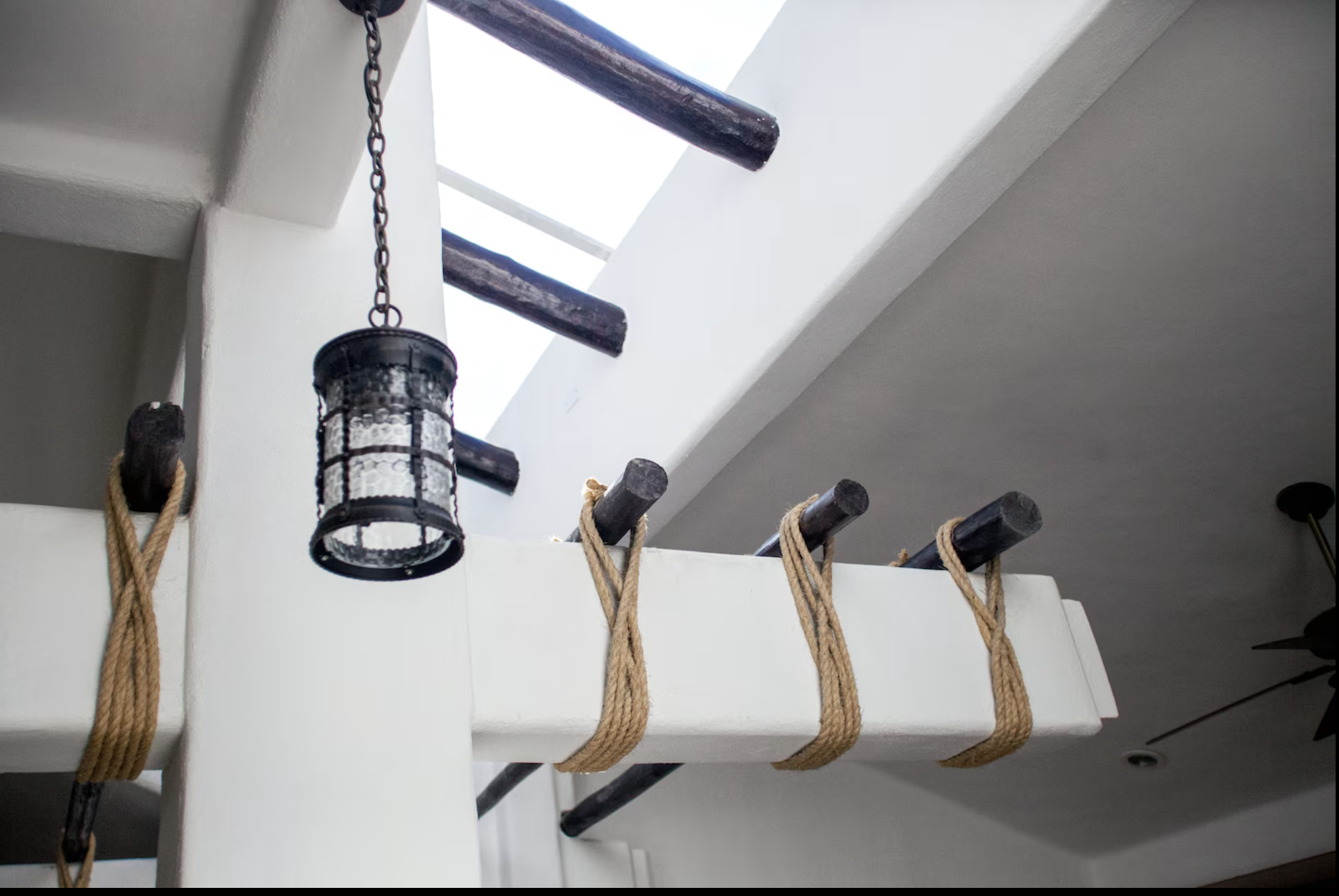 A ceiling with wooden beams, a skylight, and a hanging black metal lantern, with ropes tied around black horizontal bars.