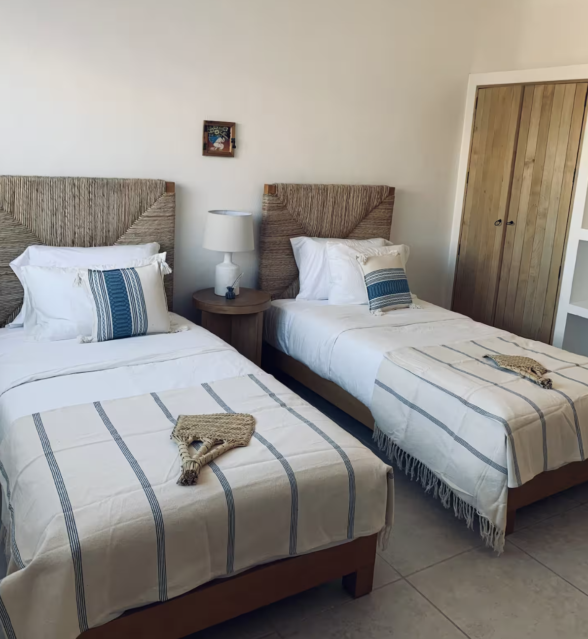 Two twin beds with white bedding and blue-striped pillows, a wooden nightstand with a table lamp and small decorative item, a closet with wooden doors, a small framed picture on the wall, and a tiled floor.