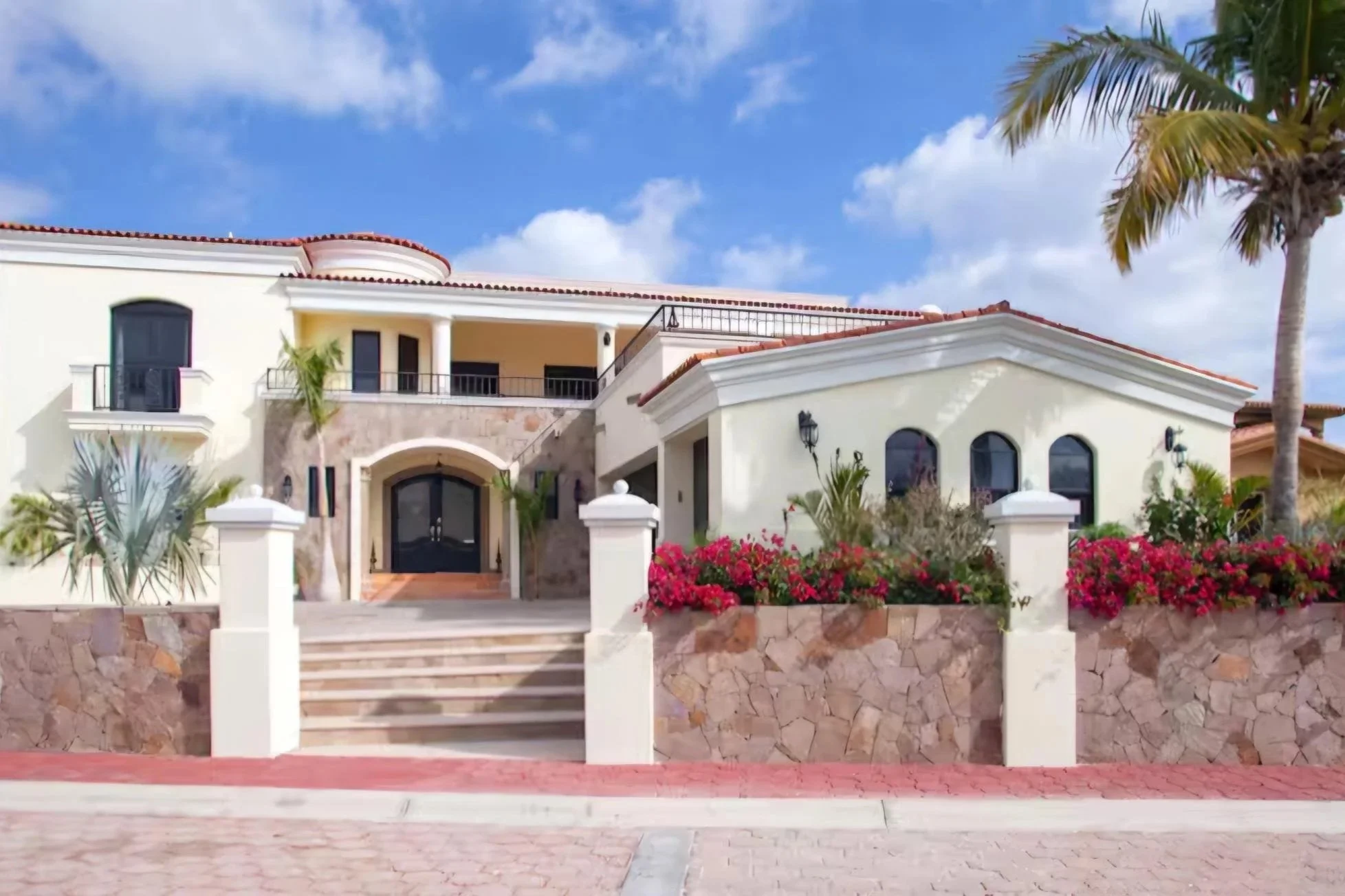Front view of a large, luxurious house with a white exterior, red-tiled roof, stone accents, and an arched entranceway. The house is surrounded by pink flowers, palm trees, and a stone wall, under a blue sky with clouds.