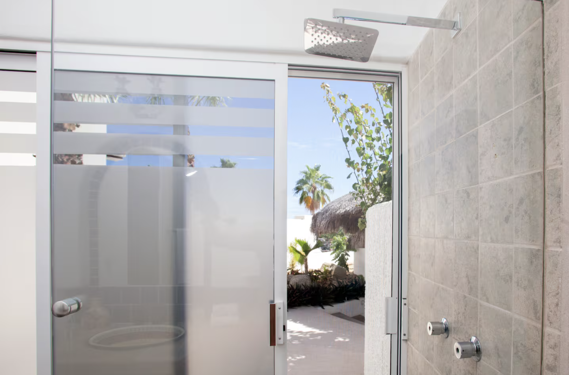 An outdoor shower with a rain shower head on the ceiling, facing glass doors that lead outside to a tropical garden with palm trees and thatched roof structures.