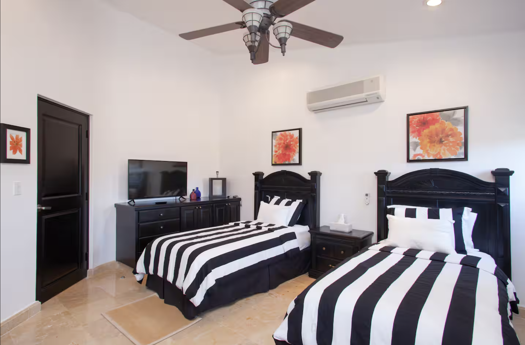A bedroom with two beds featuring black and white striped bedding, dark wooden headboards, a nightstand with tissue box, a black dresser with a TV, and framed orange floral artwork on white walls. A ceiling fan with light fixtures and an air conditio