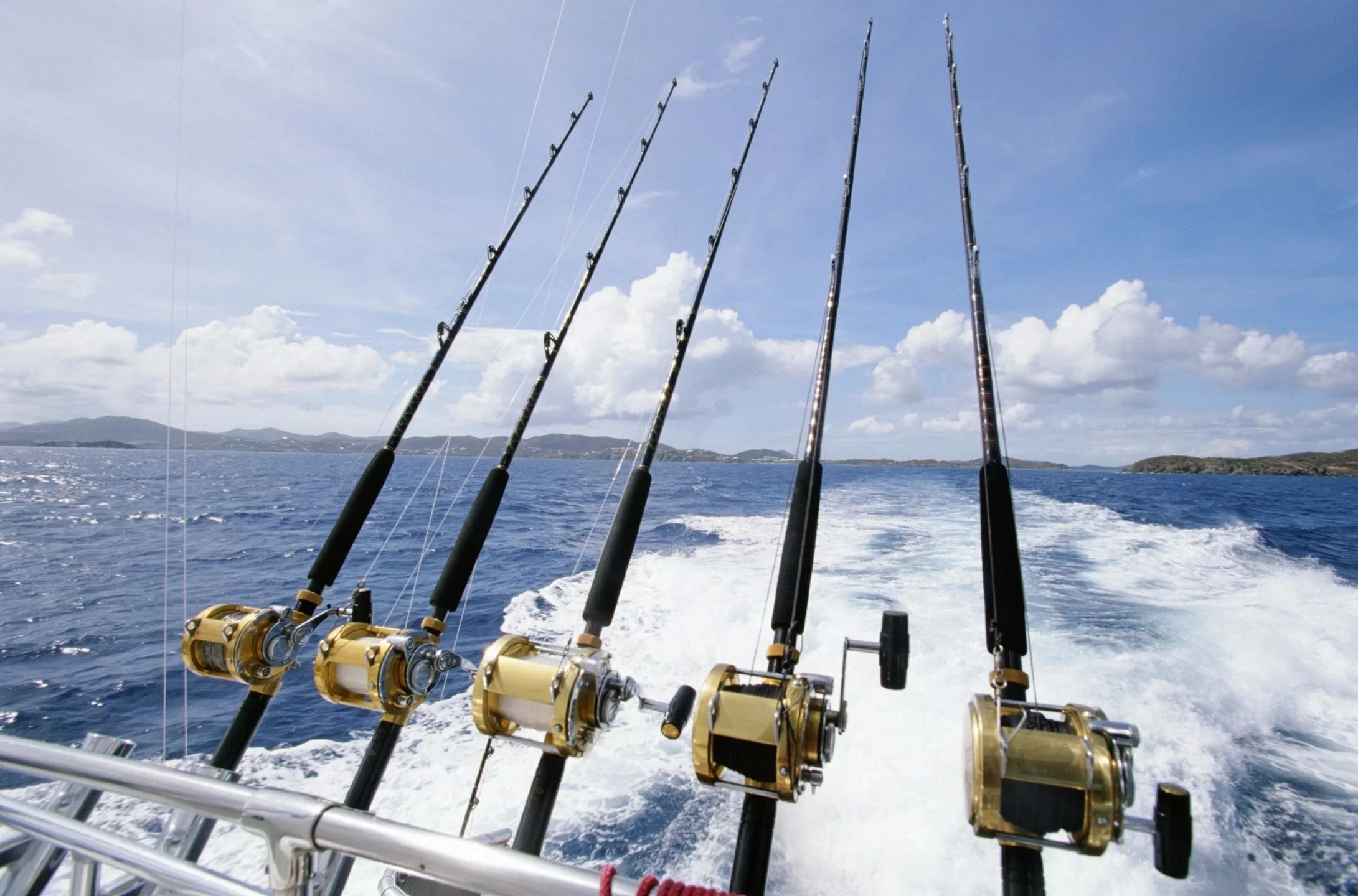 Five fishing rods mounted on the back of a boat with a wake behind, serated islands in the background and a partly cloudy sky. The Marlin Hunter fishing charter deep sea fishing