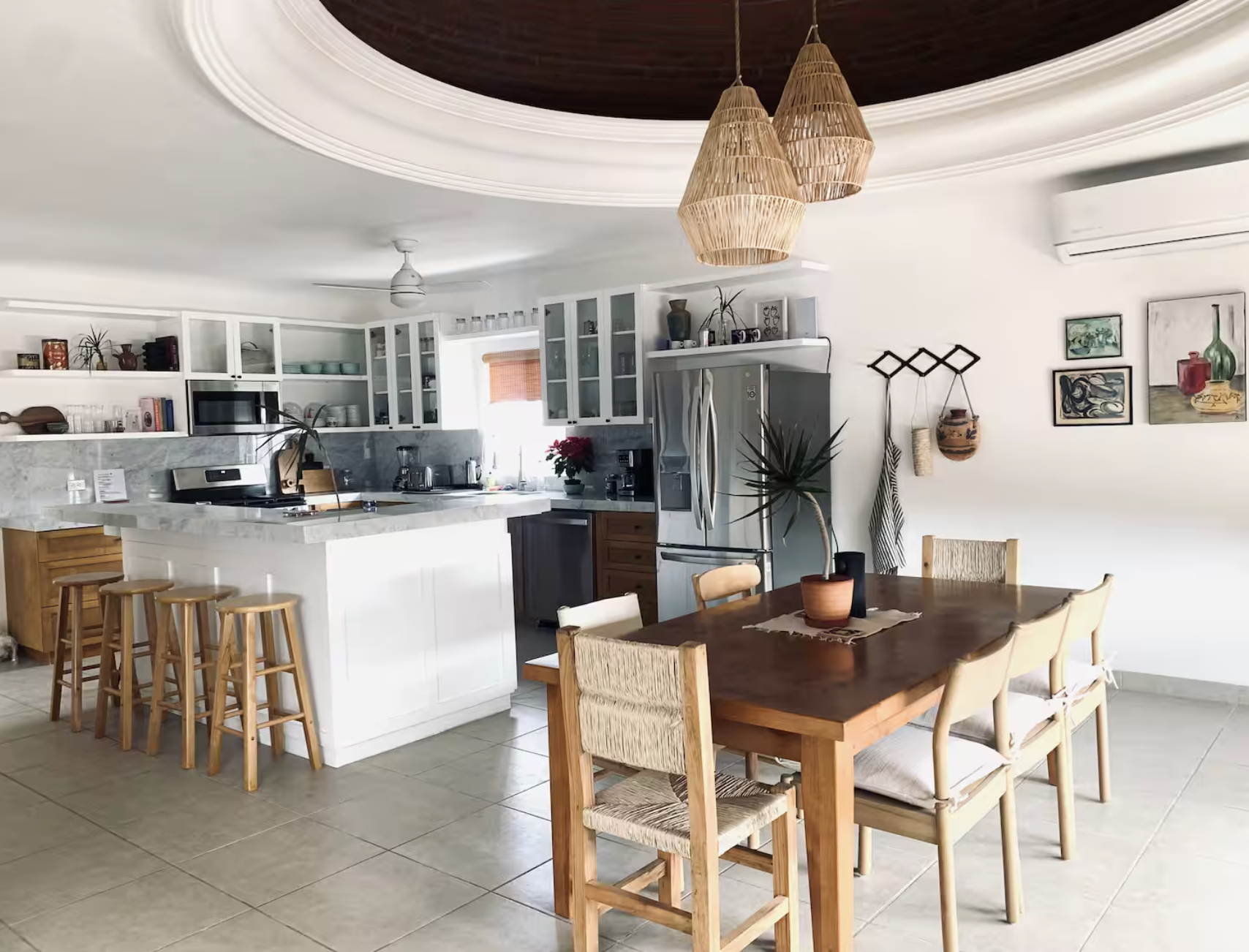 A modern open-concept kitchen and dining area with white cabinets, marble countertops, a stainless steel refrigerator, and a wooden dining table with six chairs. Decorative artwork hangs on the wall, and pendant lights hang from a tray ceiling. A pot