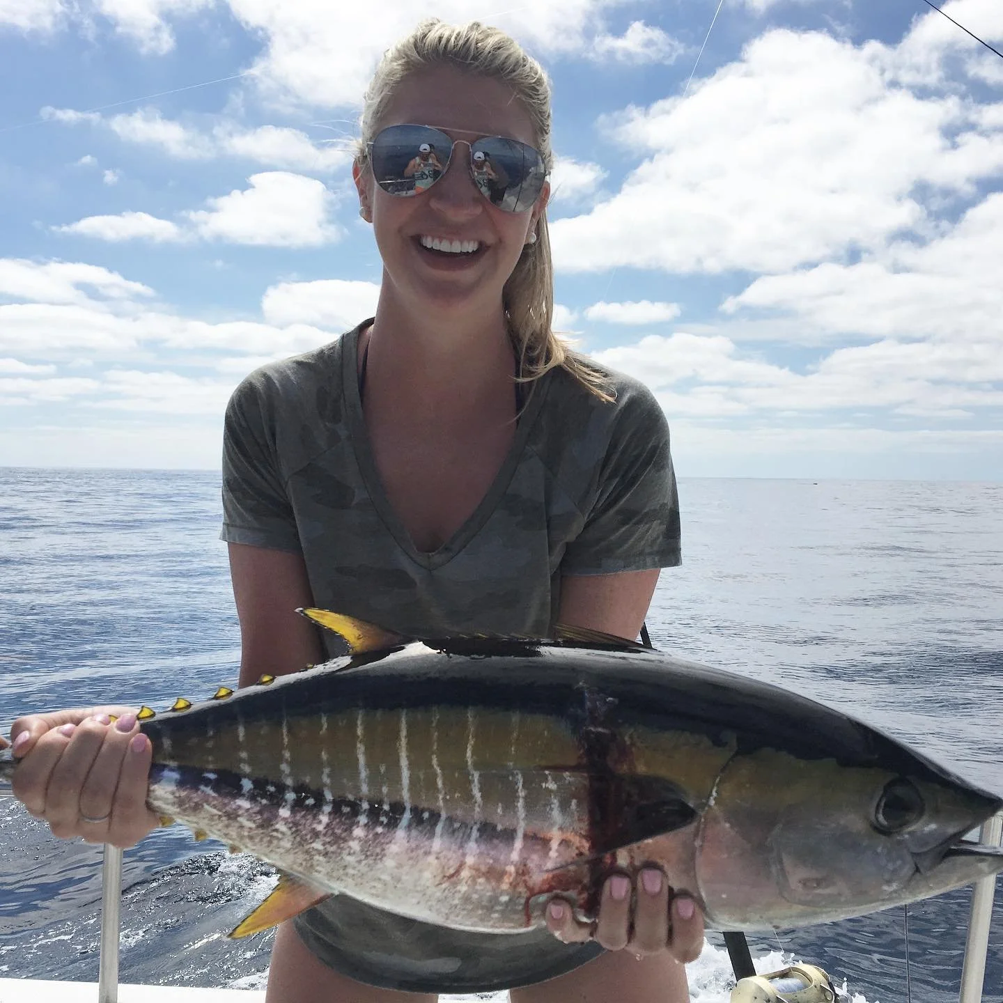 A woman in sunglasses and a camouflage shirt smiling while holding a large fish on a boat with a cloudy sky and ocean background. The Marlin Hunter fishing charters deep sea fishing