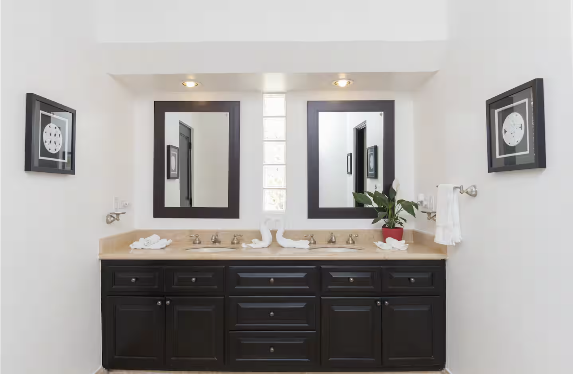 A bathroom vanity with a black cabinet, beige countertop, two sinks, and two mirrors. Two framed pictures hang on the white walls. There are towel holders, a potted plant, and towel art on the counter.