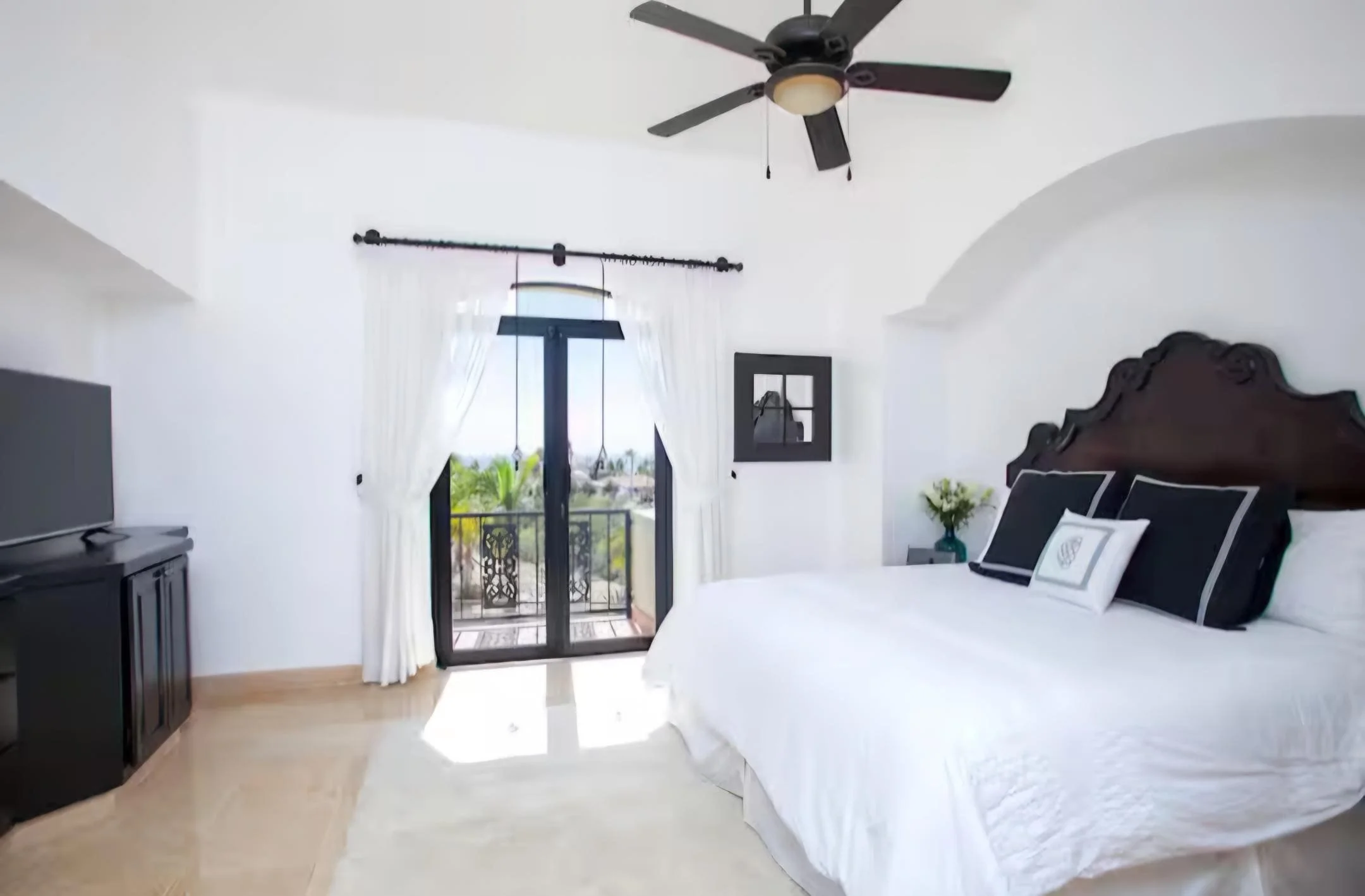 A bright bedroom with a large bed, black and white pillows, a dark wooden headboard, a small nightstand with flowers, a balcony with black wrought iron railing, and a ceiling fan. The Marlin Hunter luxury villas in Cabo