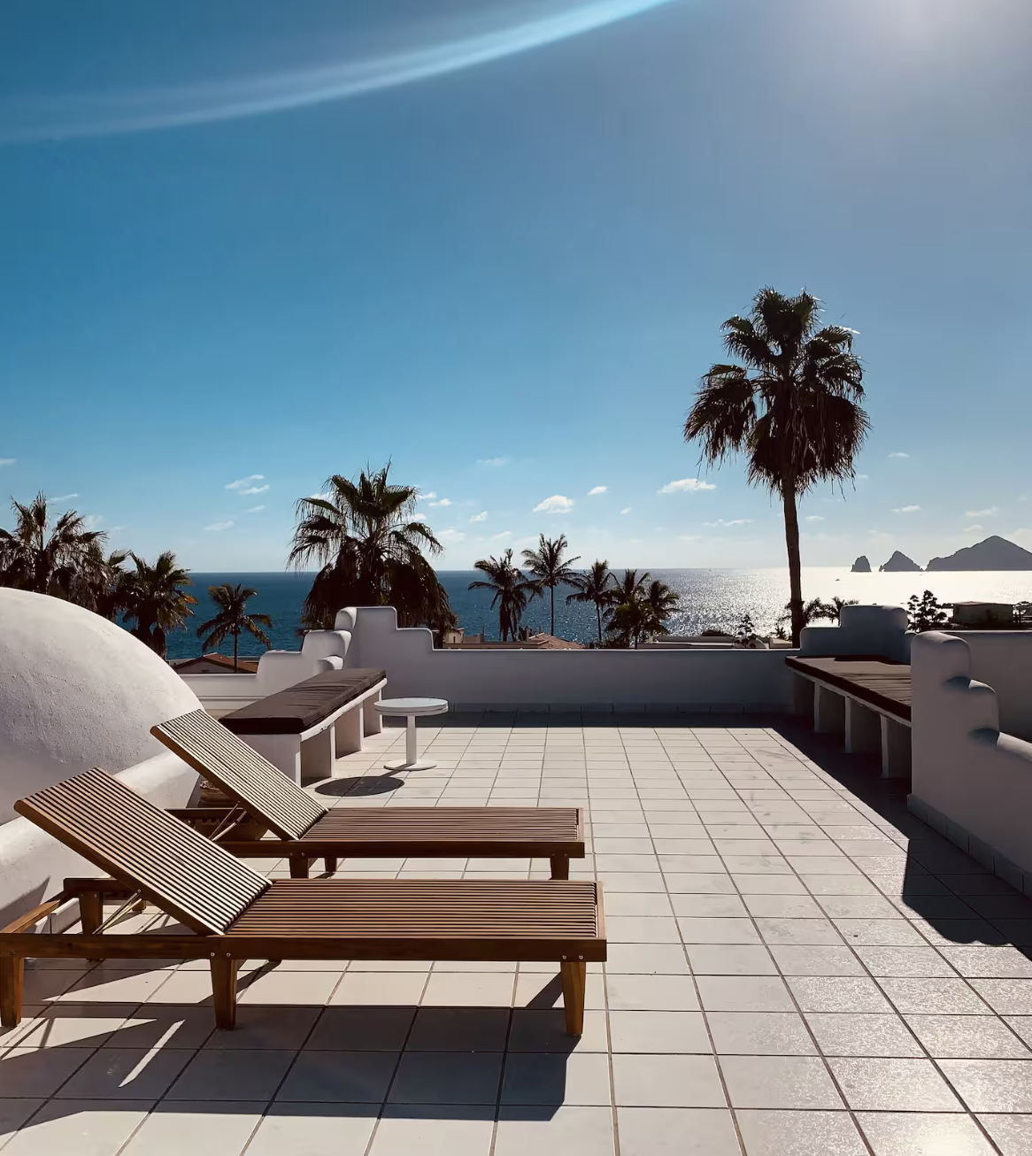 Beachside rooftop terrace with wooden lounge chairs, palm trees, ocean view, and clear blue sky