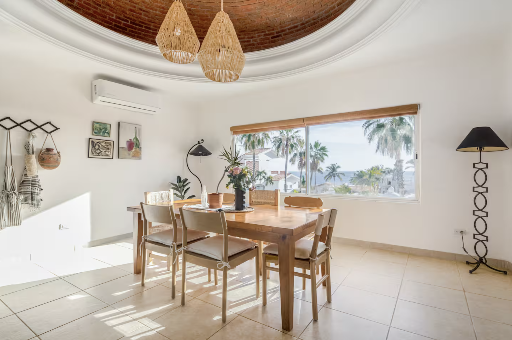 Sunlit dining room with a wooden table and six chairs, decorative hanging lamps, wall art, potted plants, large window showing palm trees and a tropical scene.