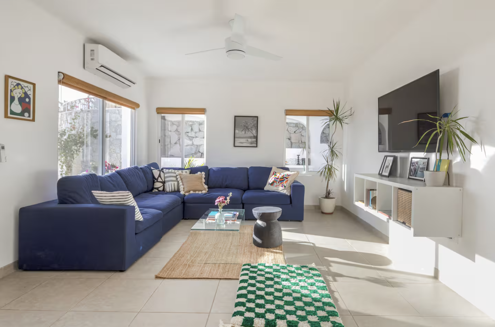 Bright living room with a large blue sectional sofa, a glass coffee table with pink flowers, a black and white side table, and a white entertainment unit with framed photos and a potted plant. Windows with blinds, a ceiling fan, and artwork on the wh