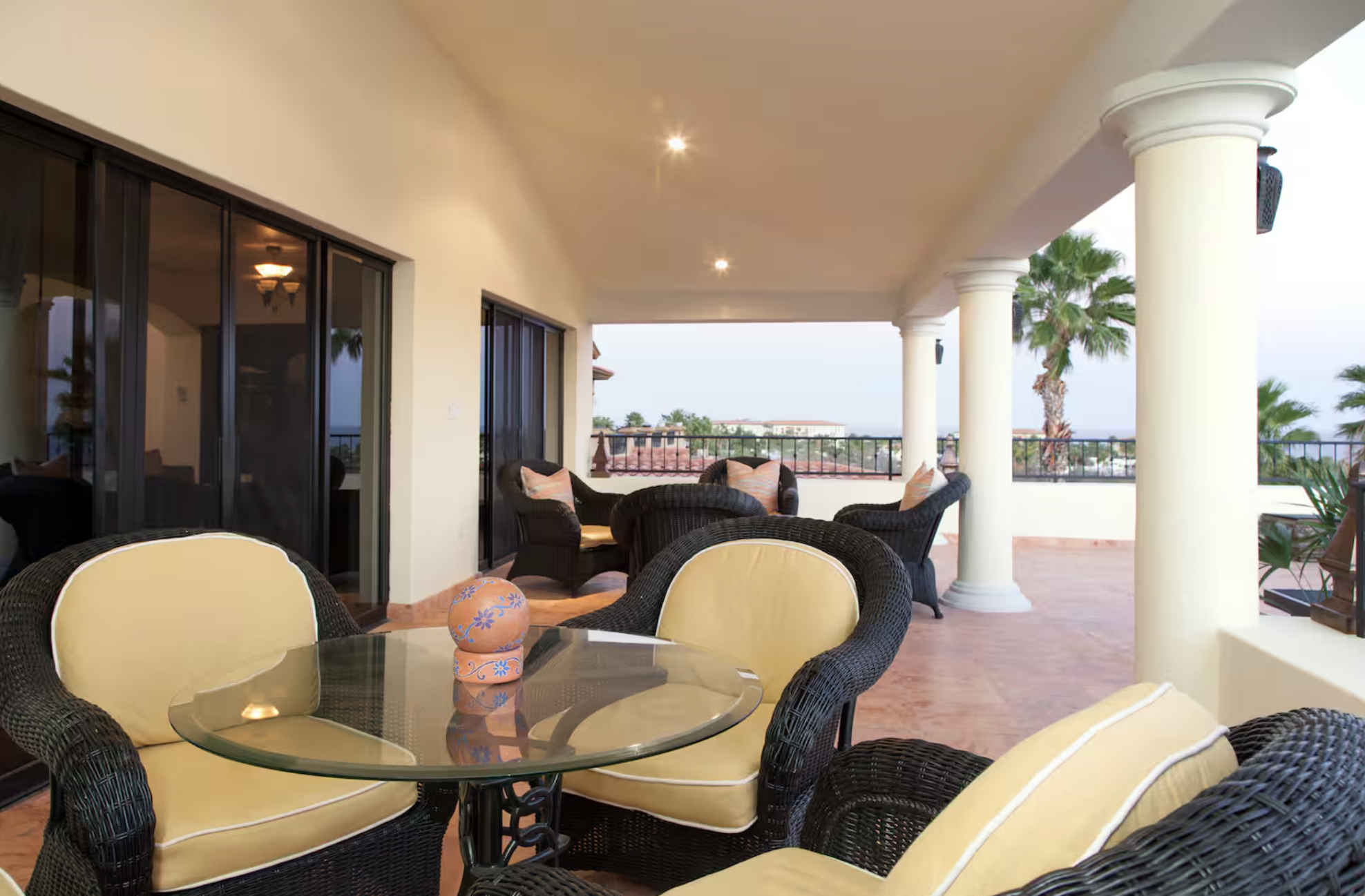 Covered balcony with wicker furniture, including a glass-top table with a decorative container, and a seating area with chairs and cushions. The balcony has columns, a tiled floor, and views of palm trees and rooftops in the background.