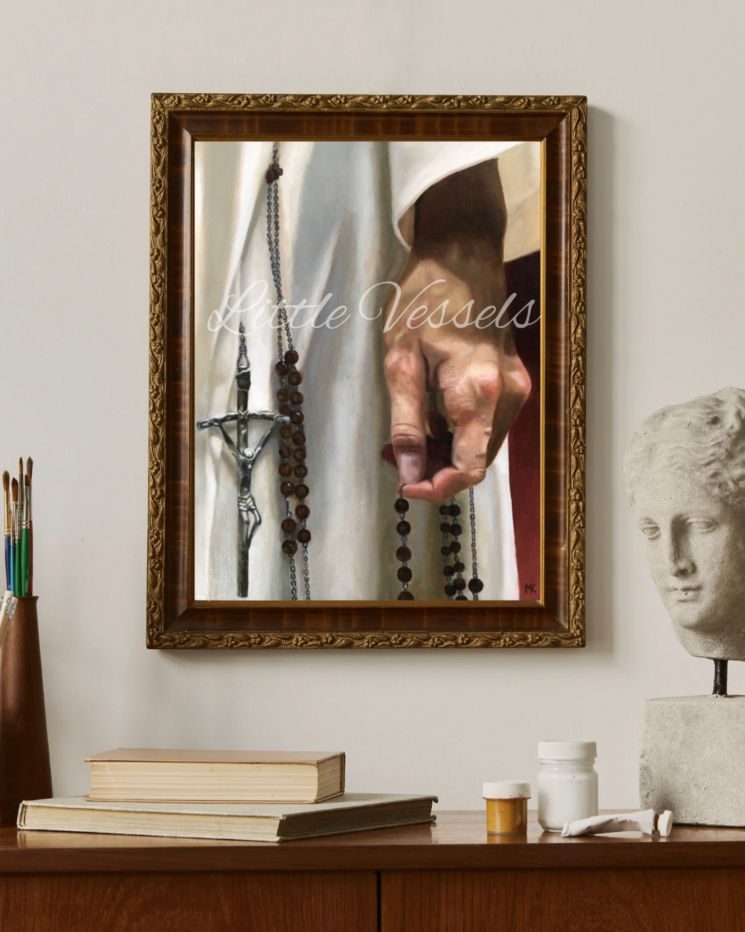 A framed painting of a man holding prayer beads, with a crucifix hanging on a white cloth. On a wooden surface below, there are books, paintbrushes, and various small containers, with a bust sculpture of a person's head to the right.