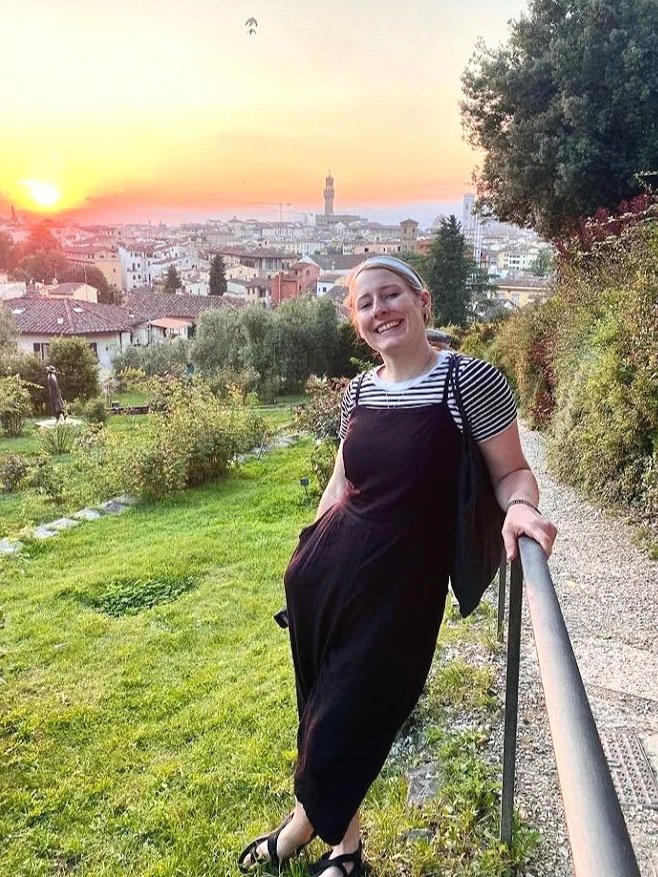A young woman with blonde hair, wearing a black jumpsuit over a striped t-shirt, holding a black bag, smiling and leaning on a railing in a park during sunset with city buildings and a tower in the background.