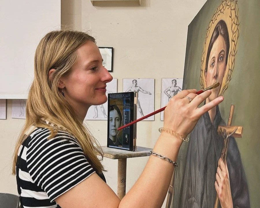 A woman painting a religious icon of a saint, with a phone on a stand displaying the subject's face.