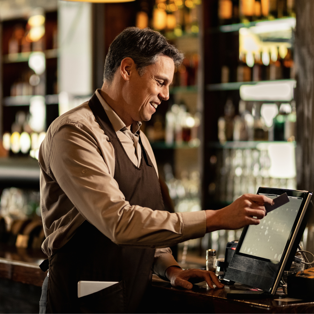 A smiling male bartender working behind the bar, using a touchscreen register, with shelves of bottles in the background.