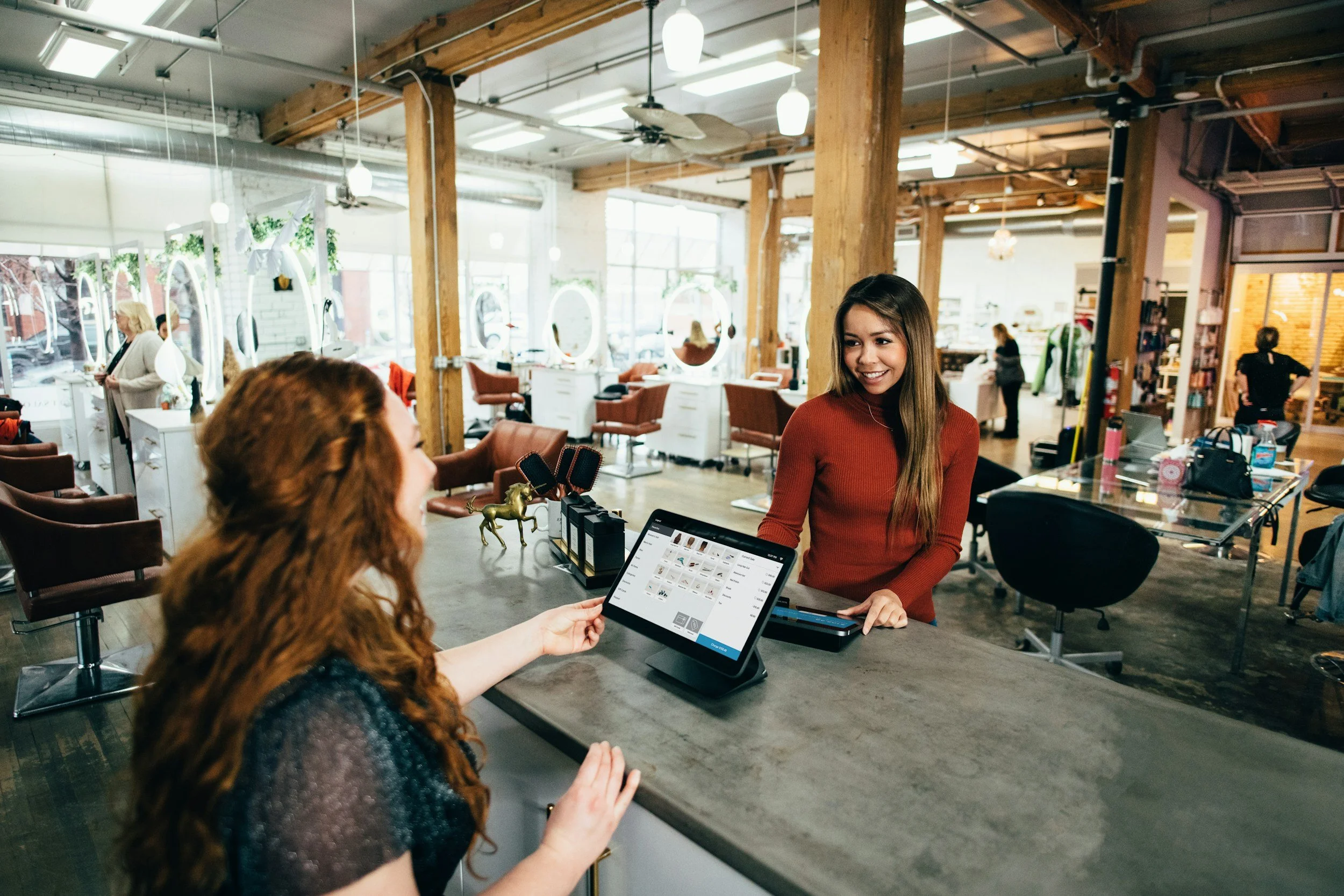 A woman with long brown hair in a red sweater smiling while speaking with another woman with curly red hair. The woman with curly hair is holding a tablet displaying a grid of images. They are inside a bright, modern shop or cafe with large windows, wooden beams, and various seating and decor.