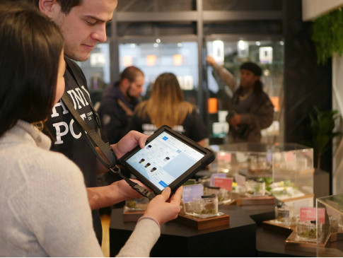 Two people look at a tablet inside a store, with a display of products behind them and a staff member assisting customers in the background.
