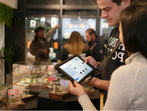 Two people look at a tablet in a store with display cases containing jewelry or small items, and other shoppers in the background.