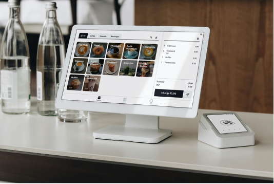A tablet displaying a digital menu with food images and prices, connected to a card reader on a white counter in a cafe or restaurant setting.