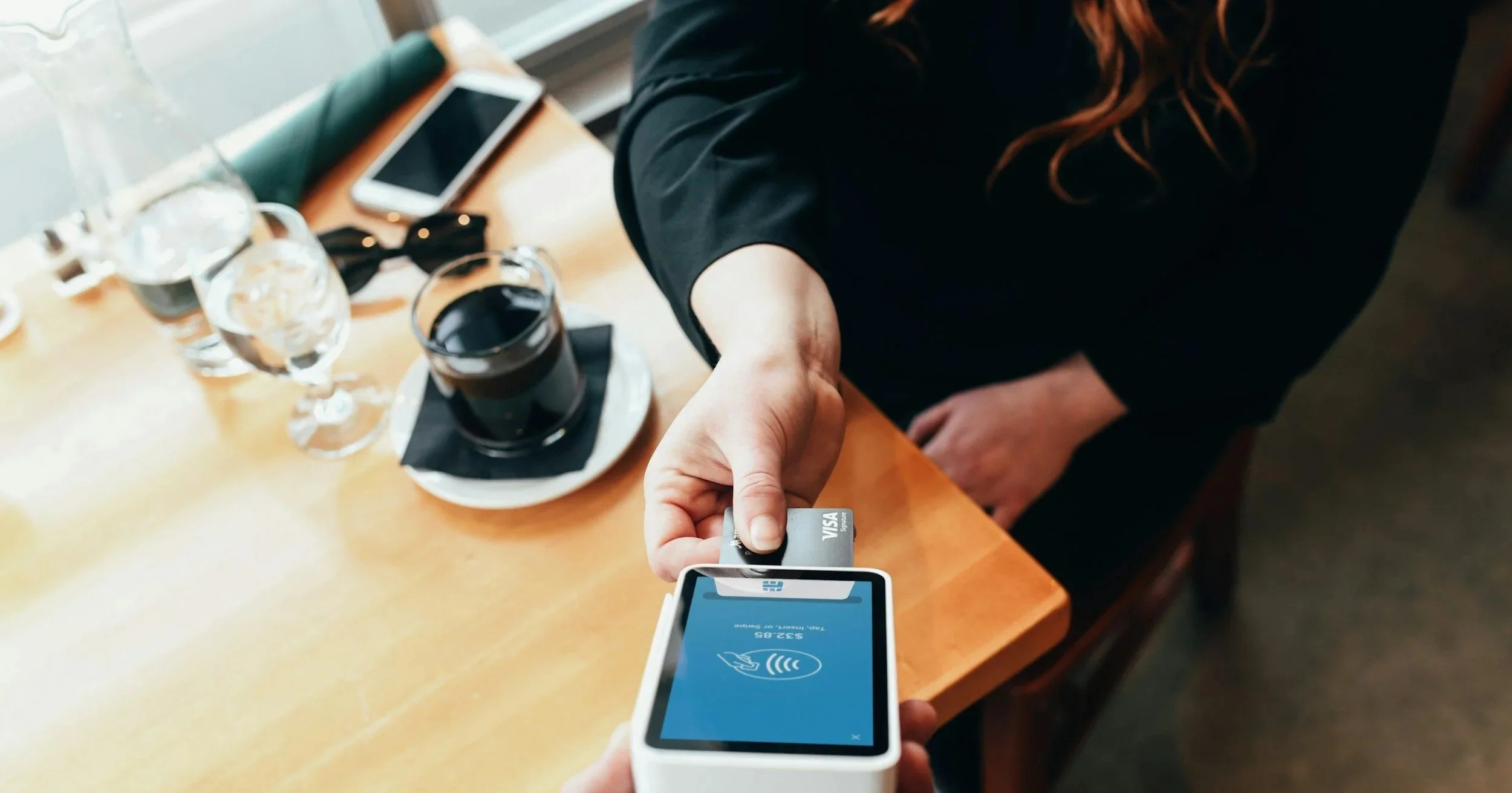 A person is paying with a contactless credit card on a mobile device at a cafe table. There are glasses of water, a glass of dark soda, a smartphone, and other items on the table.