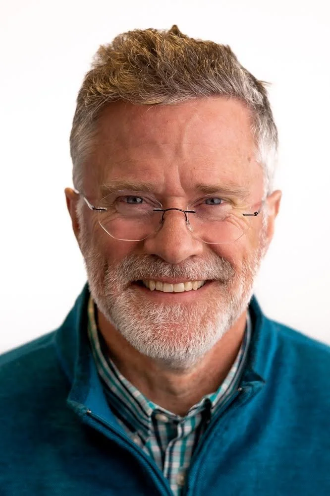 Smiling older man with gray beard and glasses, wearing a blue jacket and a plaid shirt against a white background.