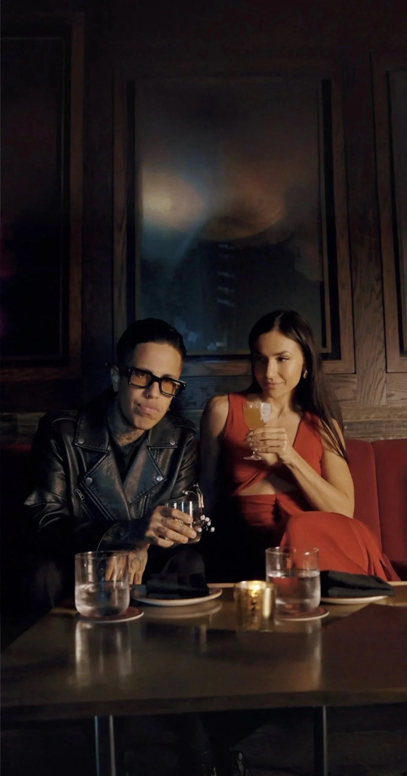 A man and a woman sitting on a red booth seat in a dimly lit bar or restaurant, holding drinks. The man is wearing glasses, a black leather jacket, and has tattoos on his hands. The woman is wearing a sleeveless red dress and has long dark hair. There are glasses of water and a candle on the table in front of them.
