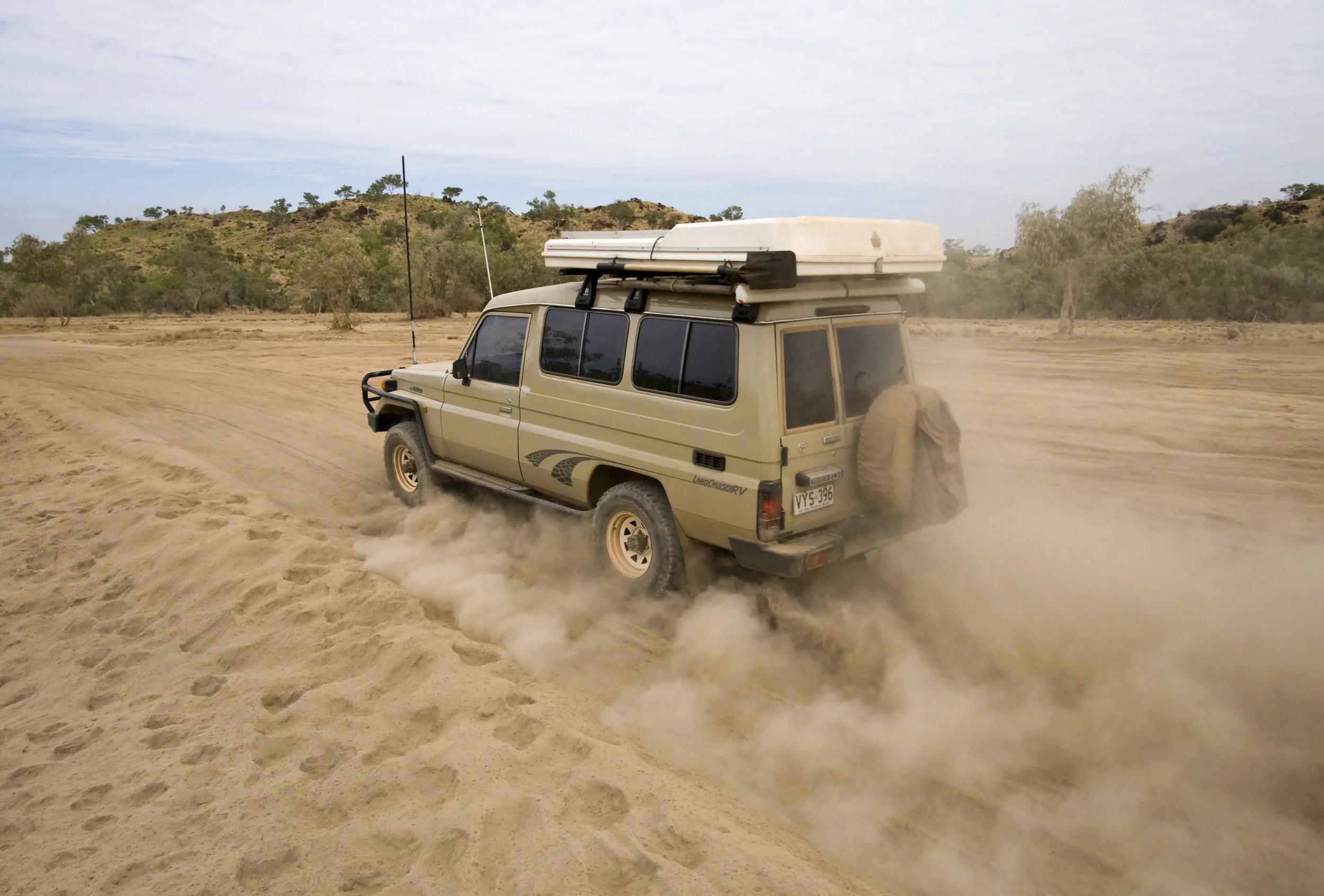 A beige off-road vehicle driving through a sandy desert, kicking up dust behind it, with a roof-mounted cargo box and a spare tire on the back.
