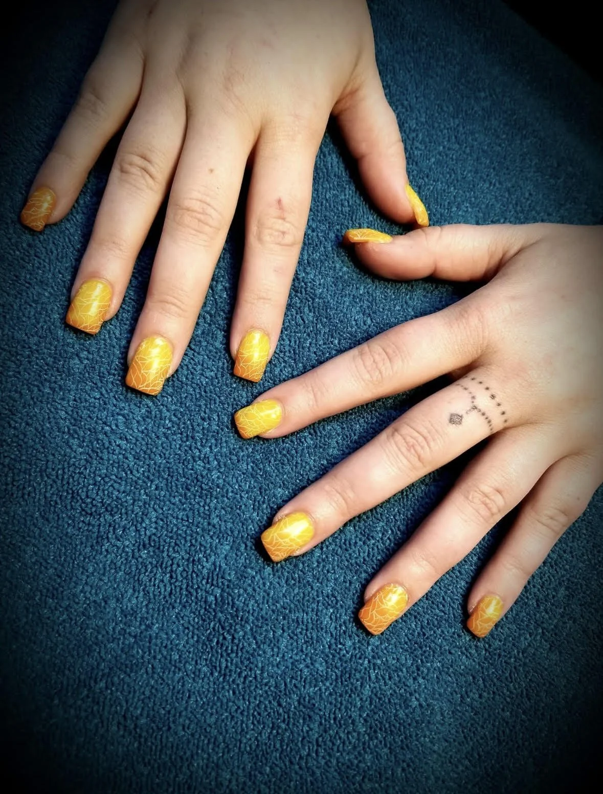 Hands with yellow-orange gradient and crackle nail polish resting on a teal textured surface. One hand has a small tattoo of a dotted line and a solid square on the index finger.