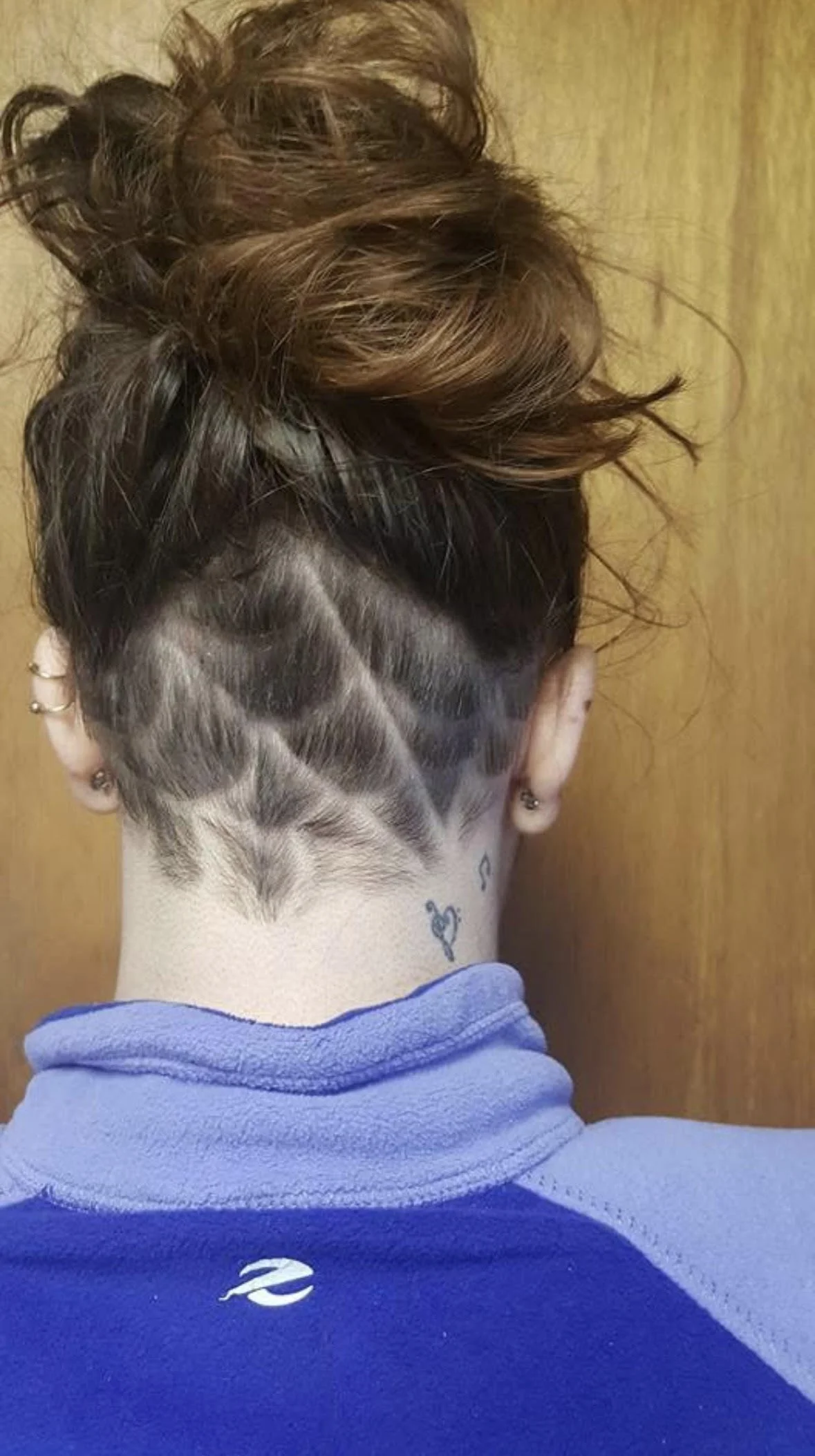 Back of a person's head with a hairstyle featuring shaved patterns at the nape and tousled brown hair on top, wearing a blue jacket with a small white logo.