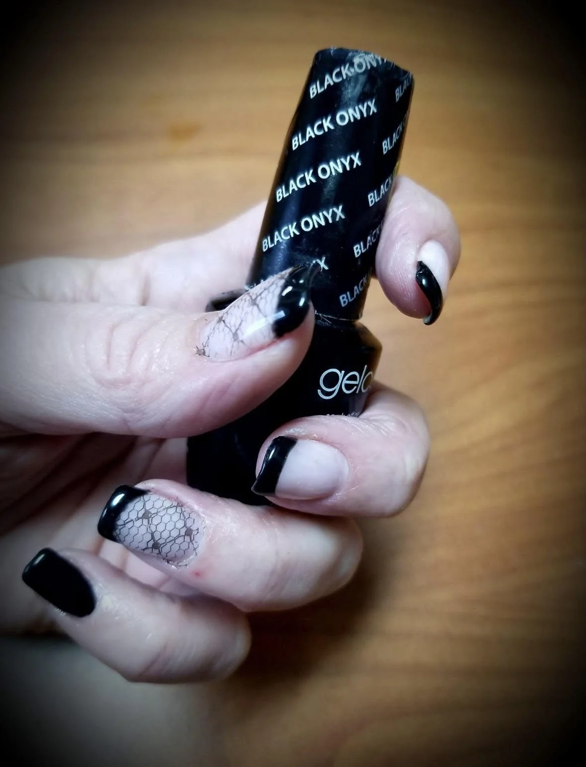 Close-up of a hand with manicured nails holding a bottle of Black Onyx gel nail polish. The nails have a black French tip design, some with a honeycomb pattern. The background is a wooden surface.