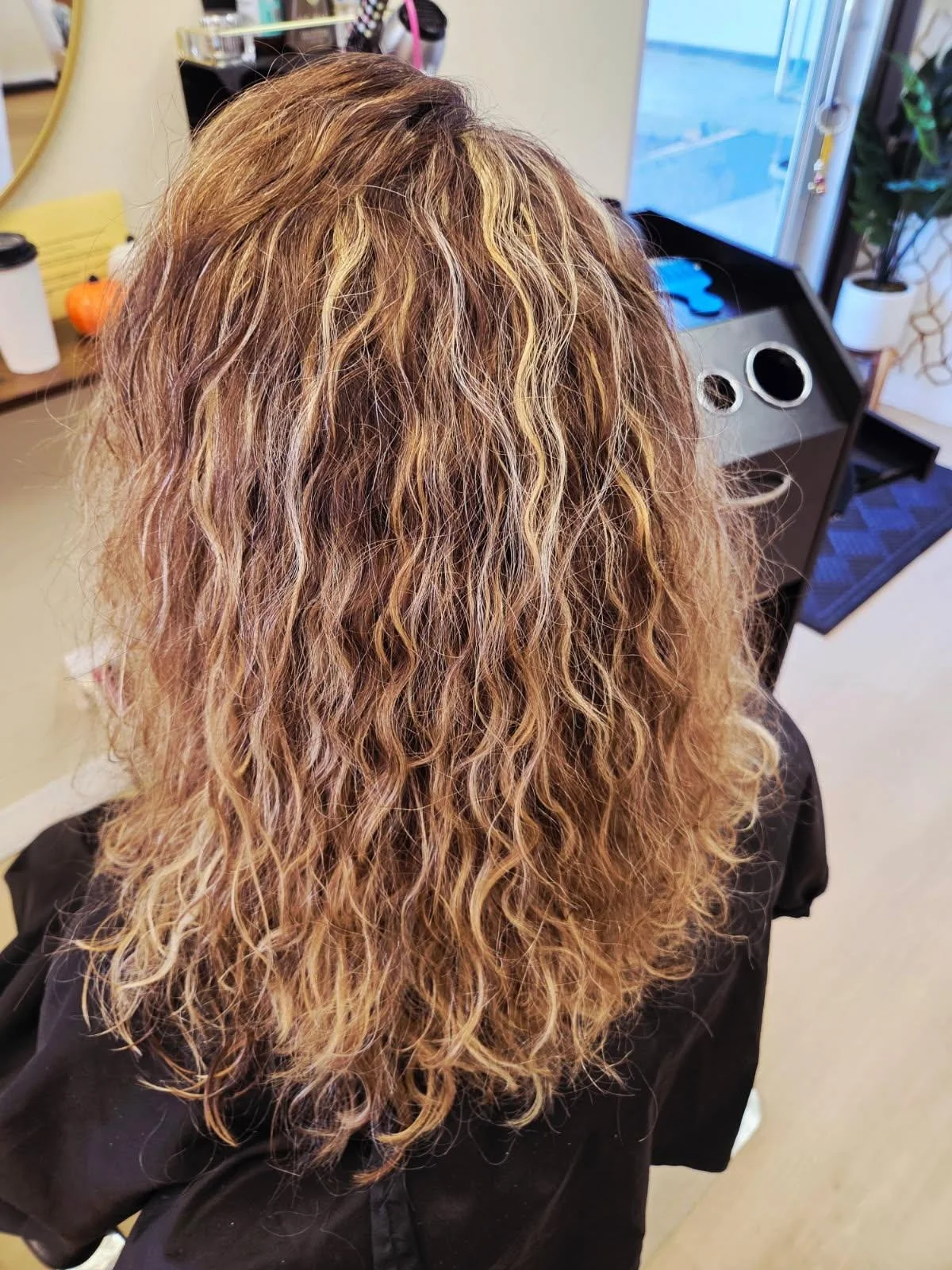 A person with shoulder-length, curly, blonde hair sitting in a hair salon.