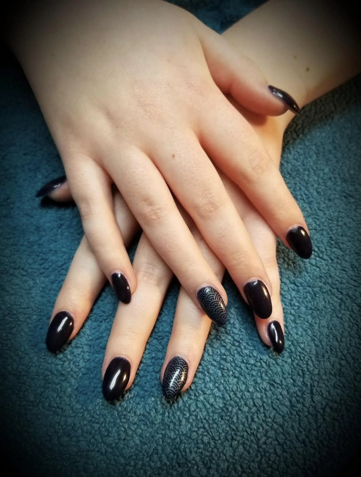 Close-up of hands with manicured nails painted dark, with decorative black and gold pattern on the ring fingernails, resting on a teal textured fabric.