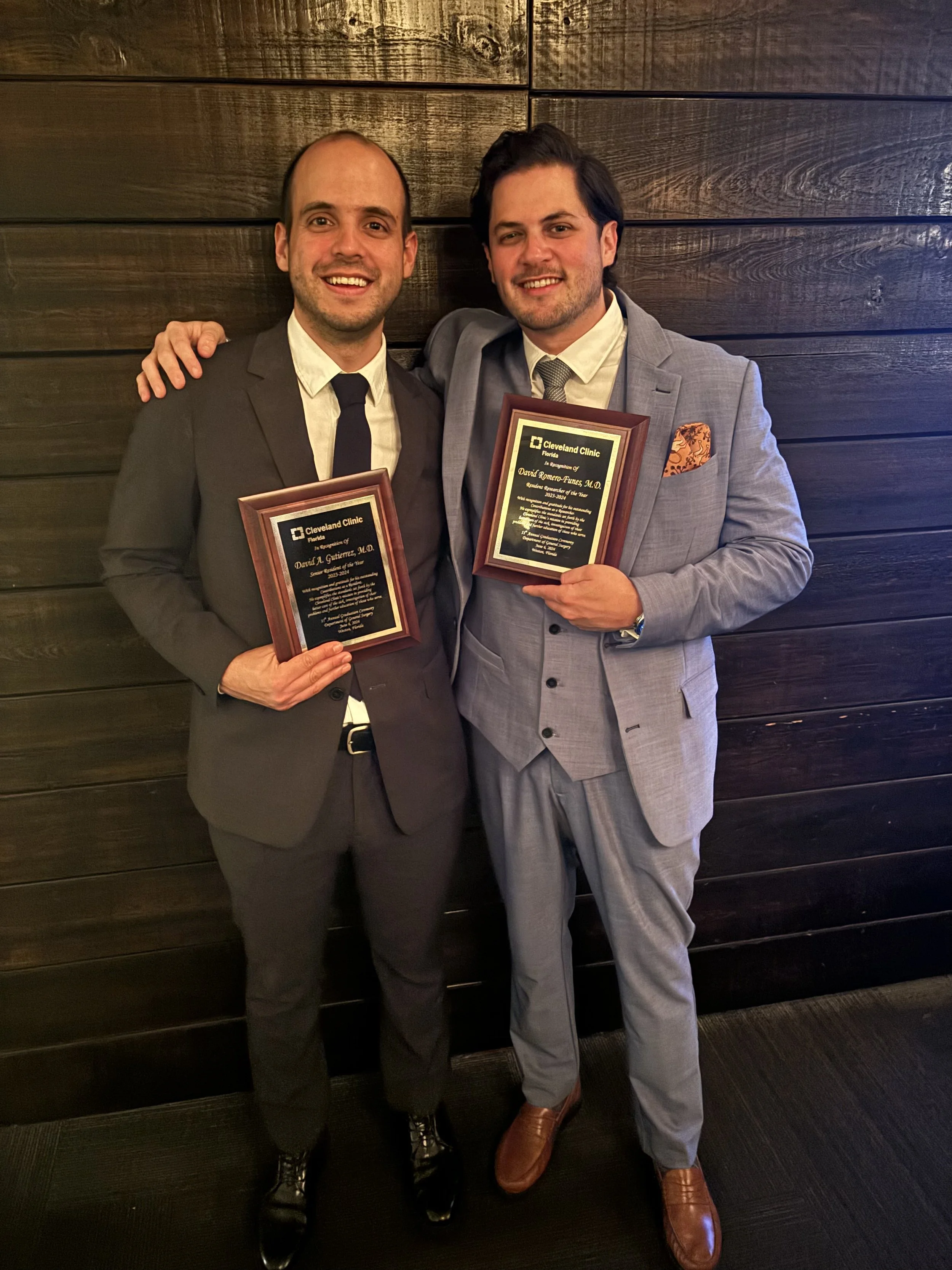 Two Surgeons  in suits holding award plaques, smiling, standing against a wooden wall.