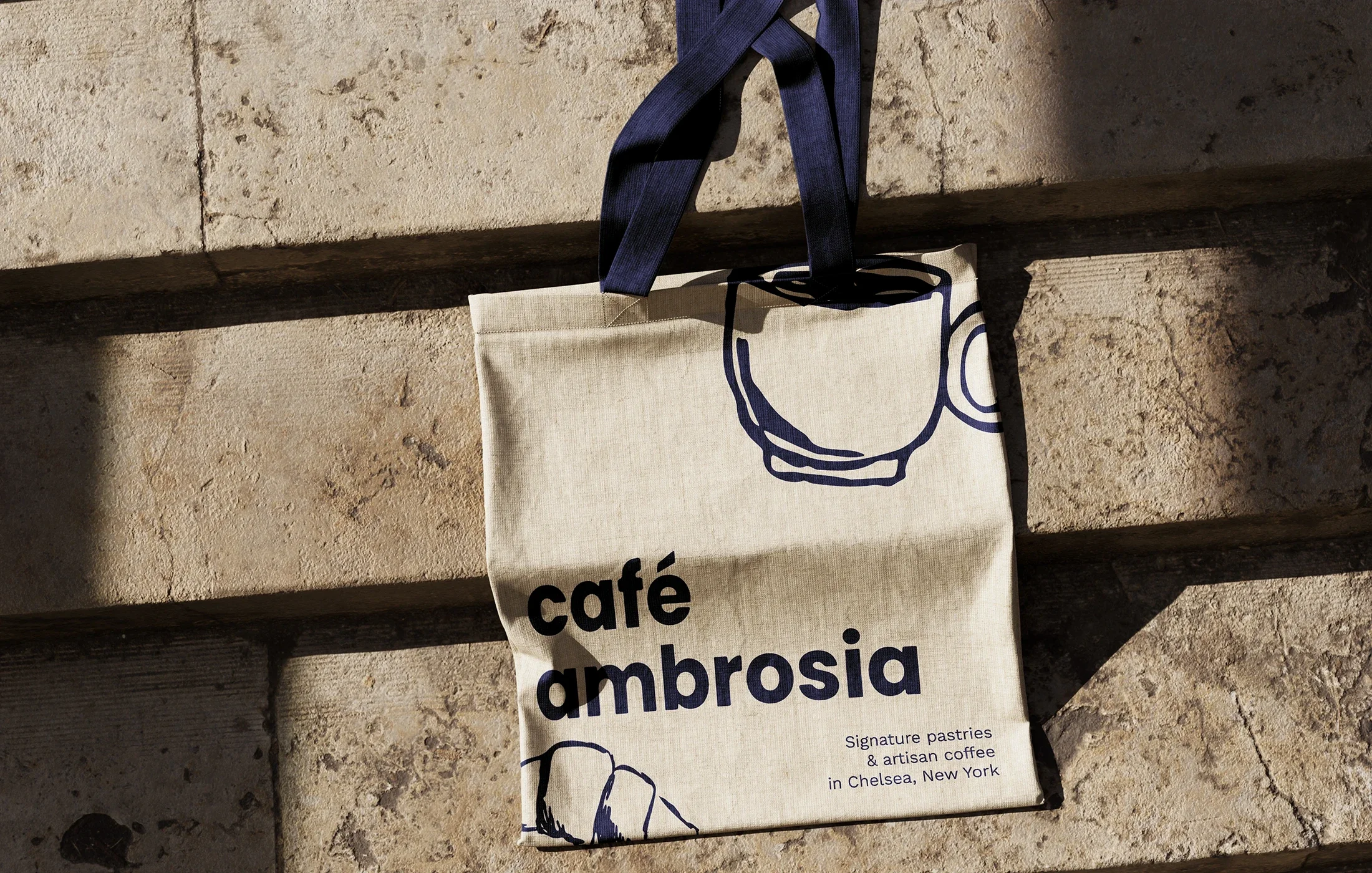 A beige tote bag with navy blue handles hanging on a stone wall. The bag has the words "café ambrosia" printed on it, along with small text about signature pastries and artisan coffee in Chelsea, New York, and sketch illustrations of a coffee cup and